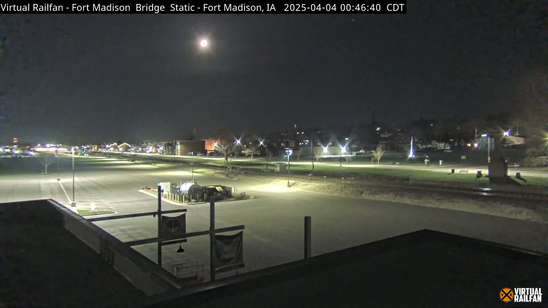 A nighttime view of a mostly empty parking lot and train tracks, with a bright moon in the sky and a few distant lights illuminating a town in the background.