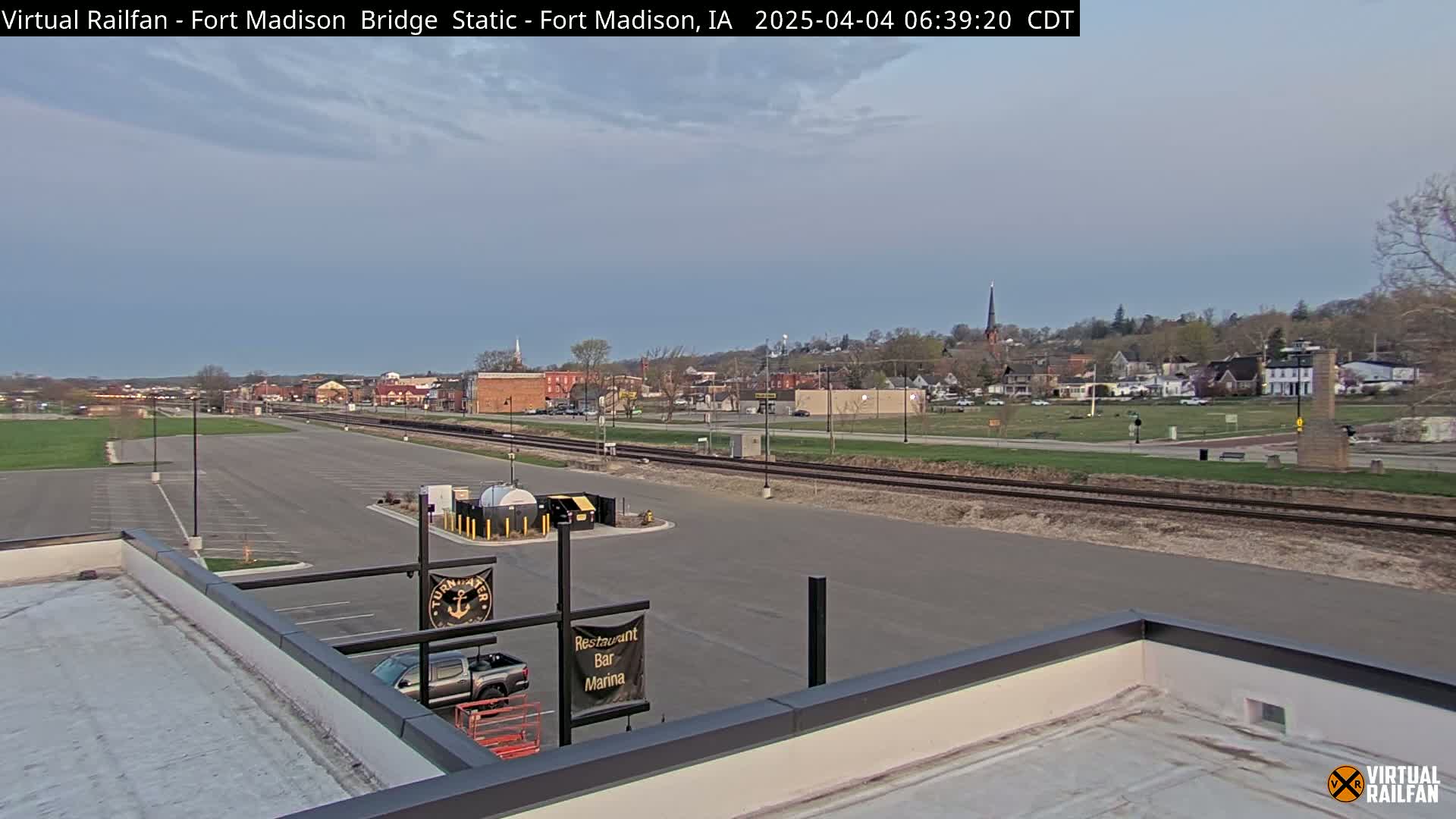 A mostly clear, early morning view of a small town with railroad tracks and a large empty parking lot, seen from a rooftop.