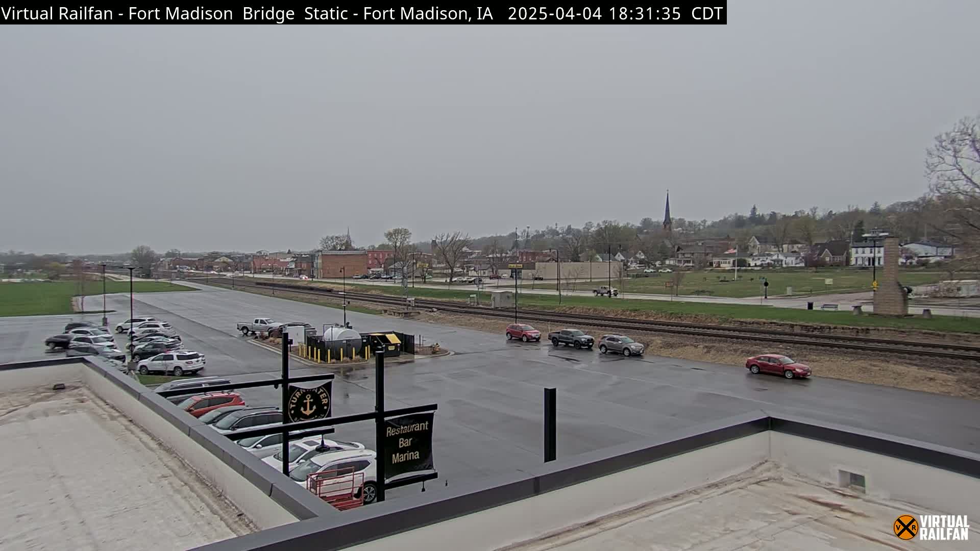 A view of a town on an overcast day, with cars parked in a lot next to a railway track, and more cars driving on a wet road.