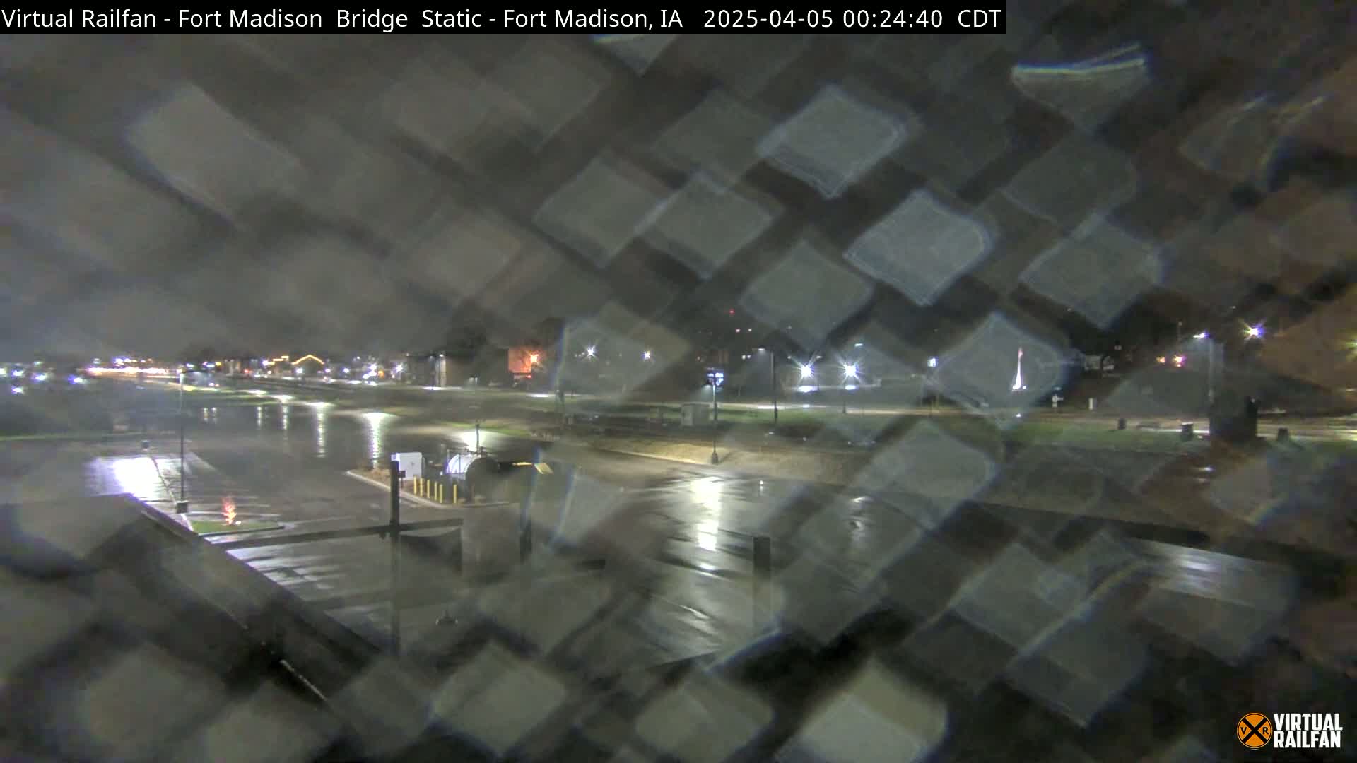 A nighttime view of a rain-slicked parking lot and roadway, illuminated by streetlights.