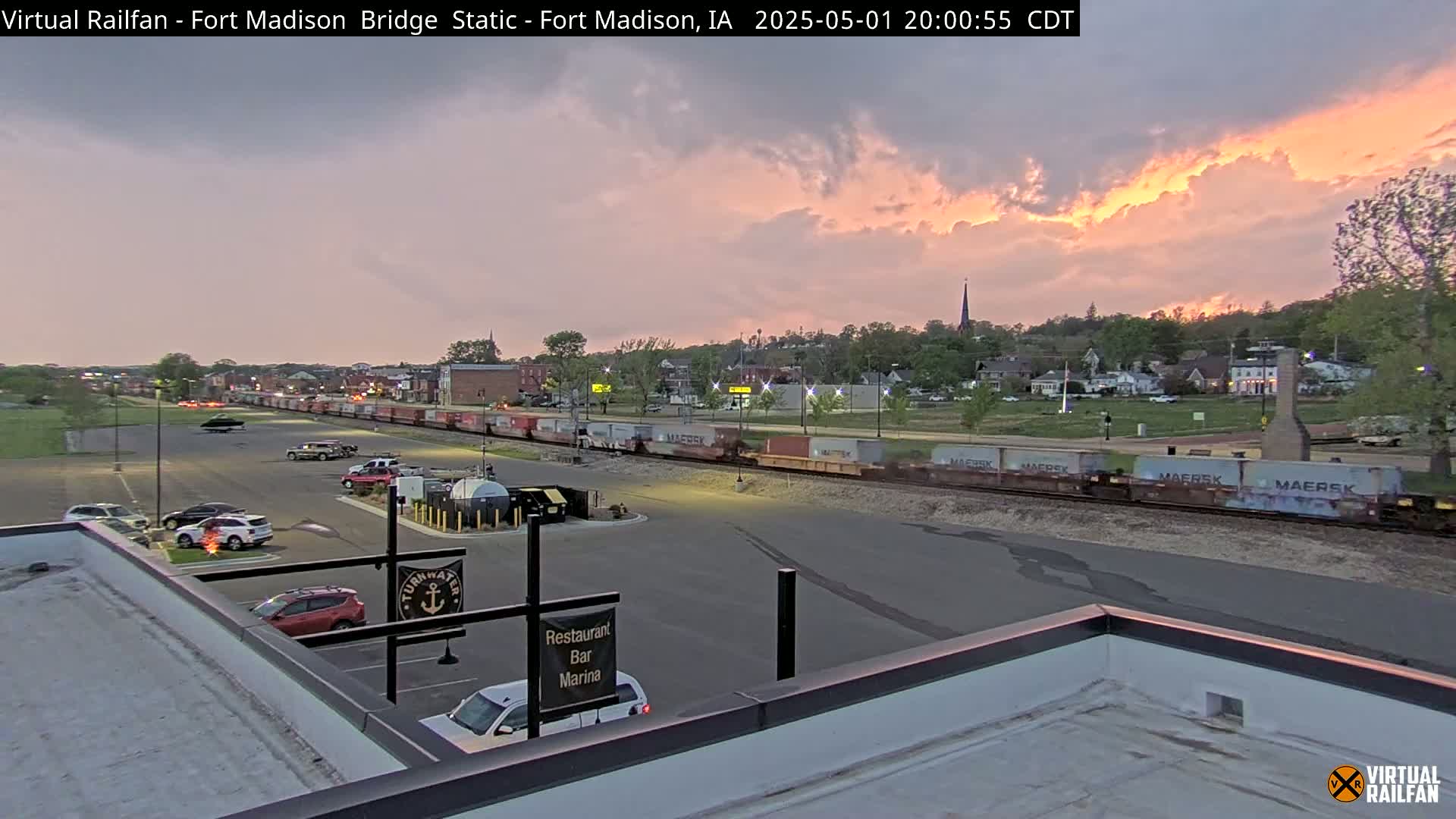 A long freight train passes through a town at sunset under a partly cloudy sky.