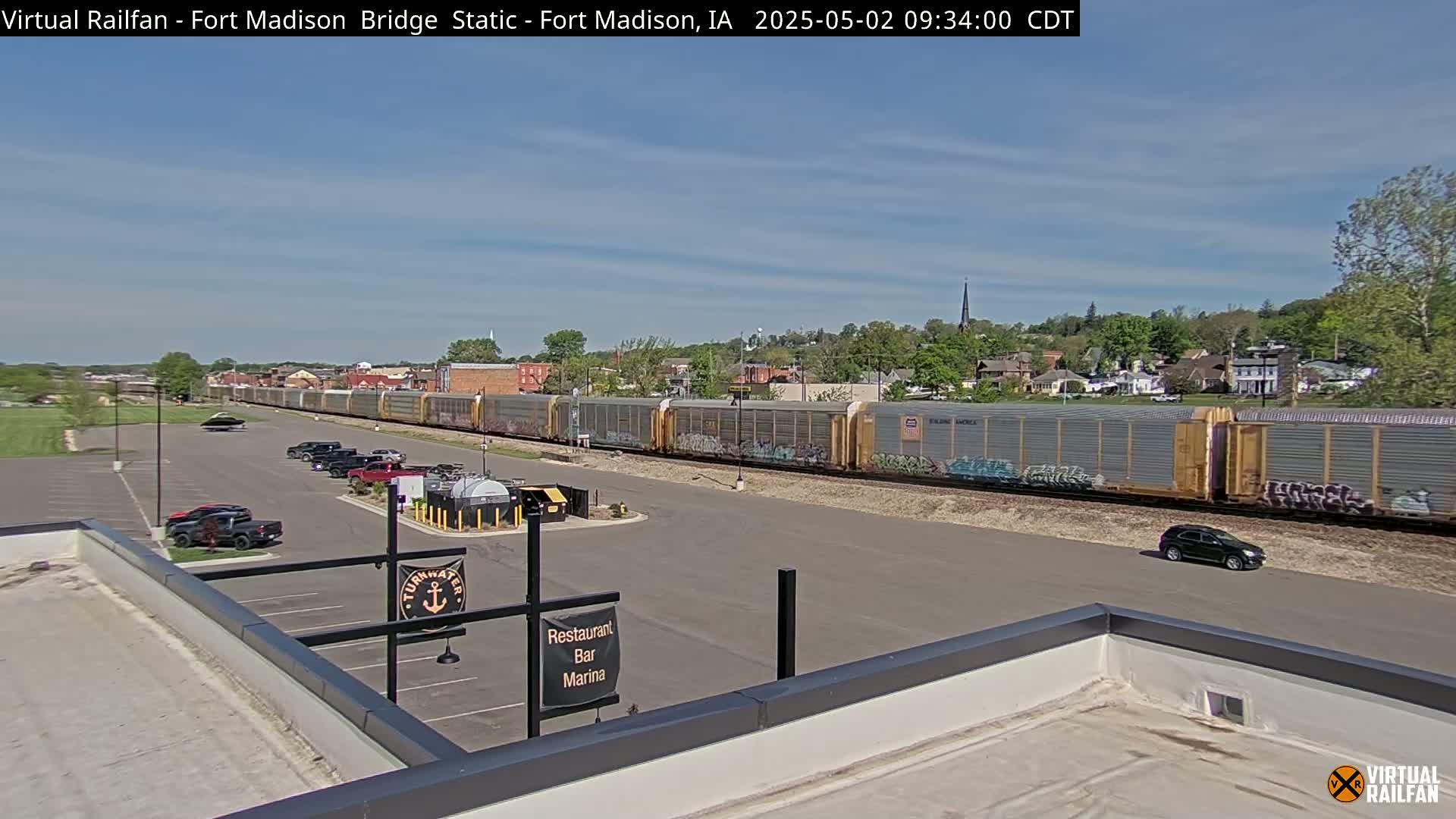 A long freight train passes through a small town under a mostly sunny sky, with a parking lot and a building visible in the foreground.