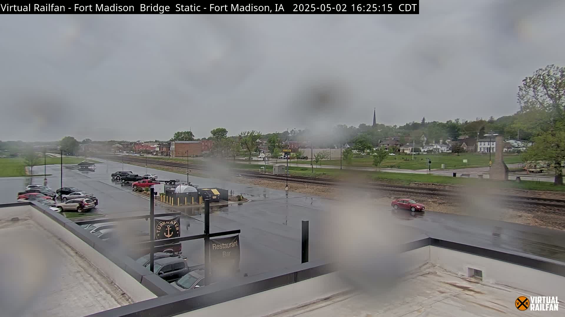 A rain-soaked view of a town's parking lot and railway tracks, with cars parked and driving, and a small town in the background.