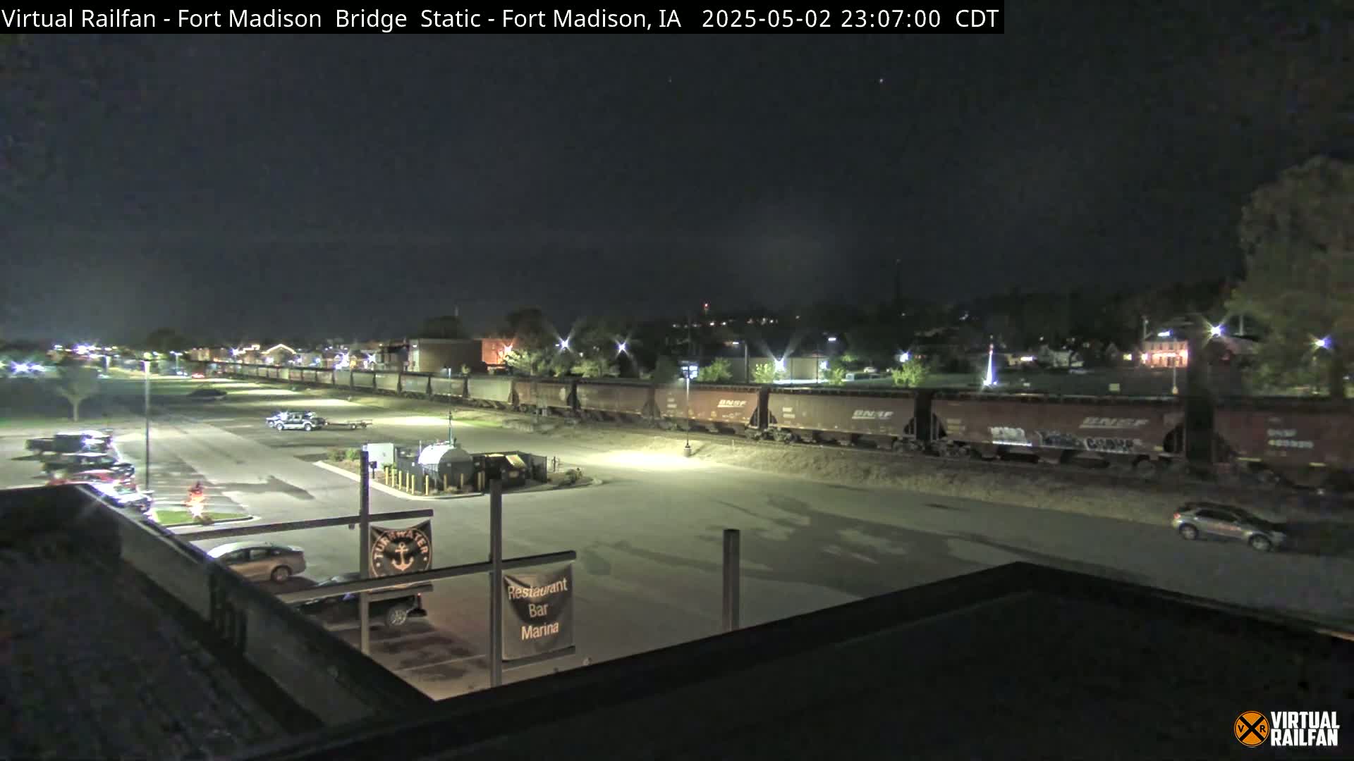 A long freight train sits stationary at night next to a mostly empty parking lot with a few cars visible, under a clear sky.