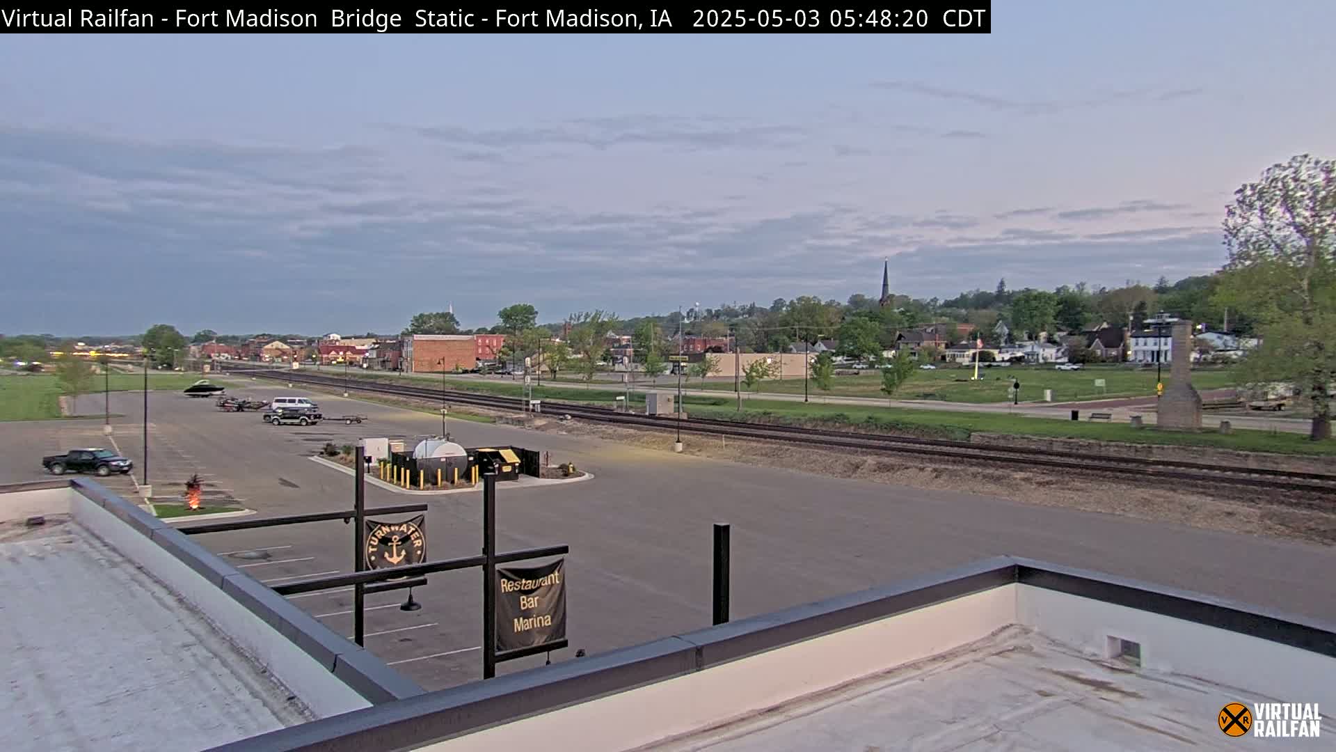 A mostly clear, twilight sky illuminates a small town's landscape, which features a parking lot, a flat rooftop, and railroad tracks running alongside a riverfront.