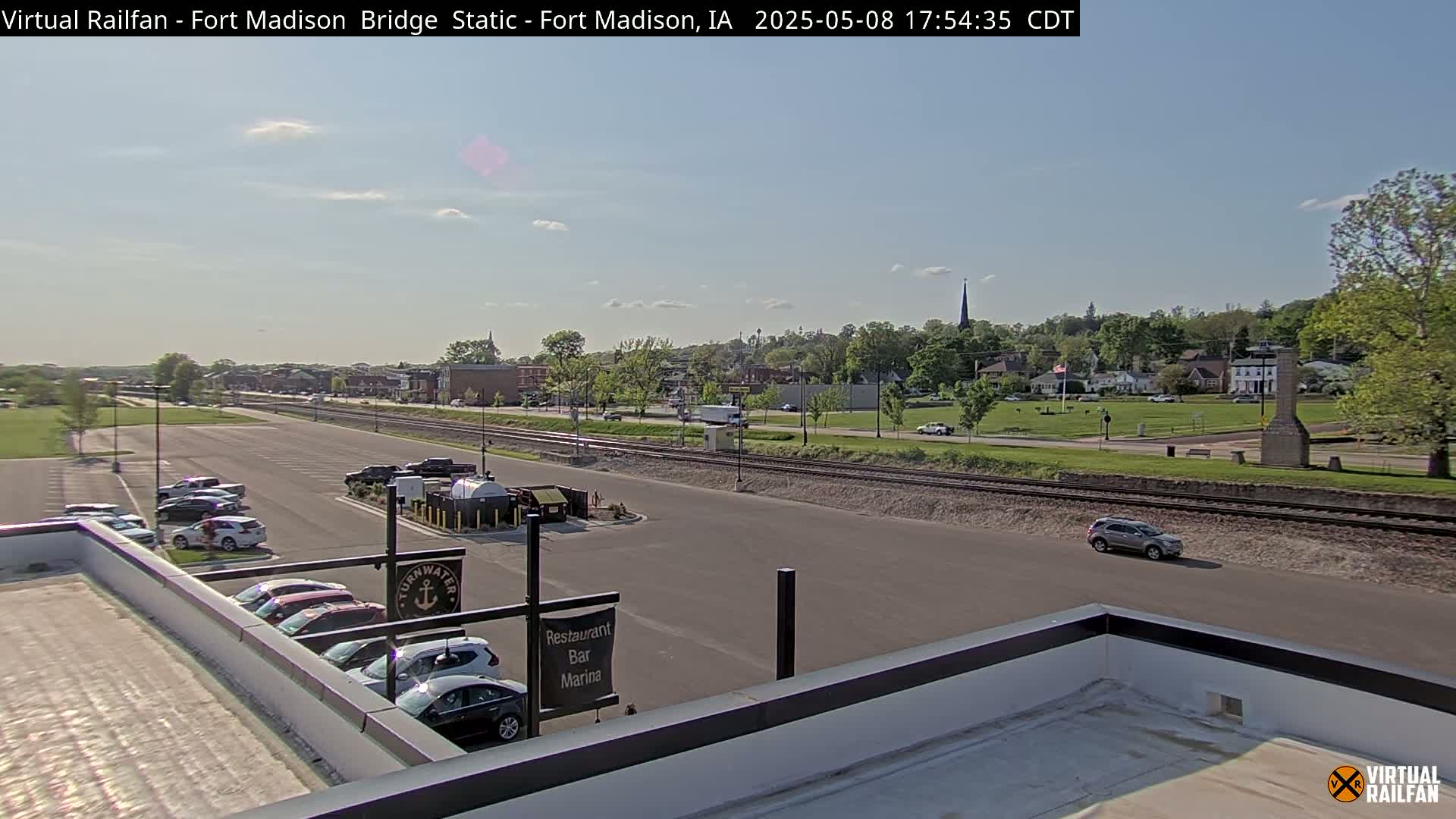 A mostly sunny view of a town with a parking lot, railroad tracks, and a few cars visible, seen from an elevated vantage point.