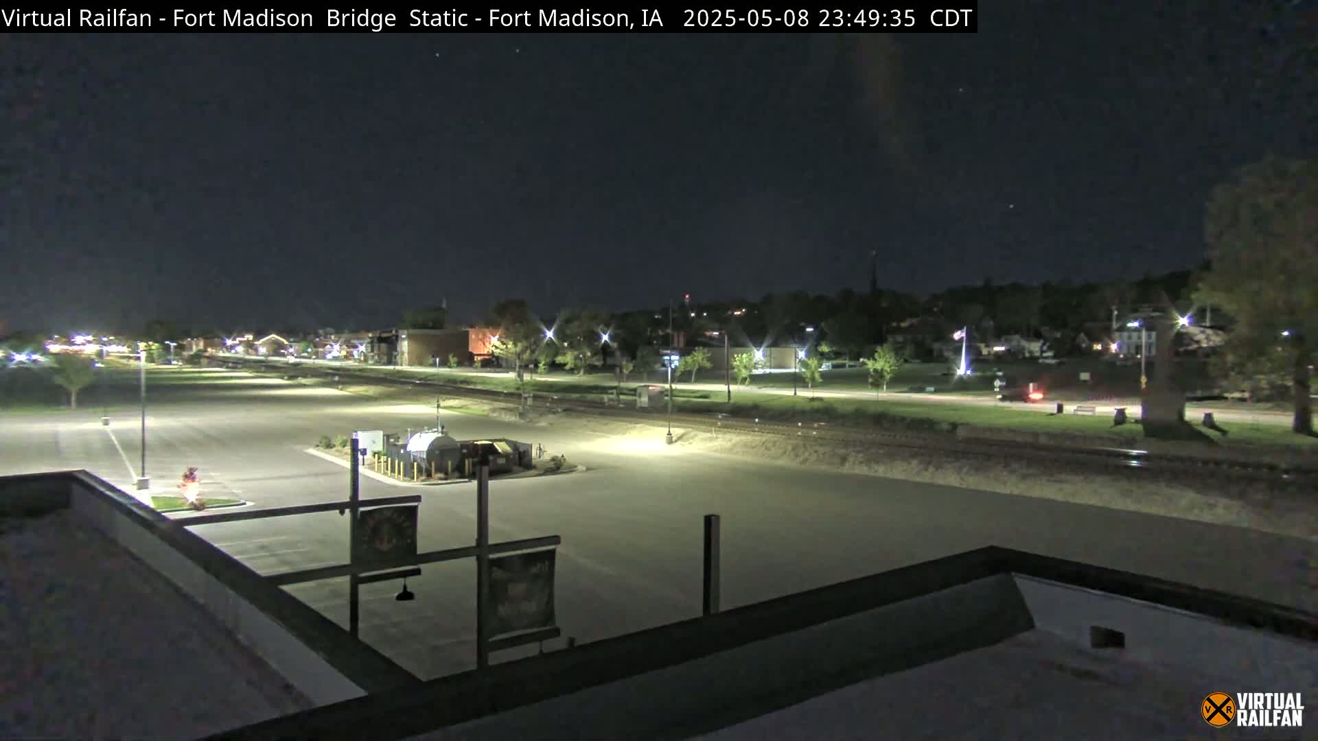 A nighttime, mostly clear view of a mostly empty parking lot and a road next to railroad tracks, with buildings and trees in the distance under nighttime lighting.