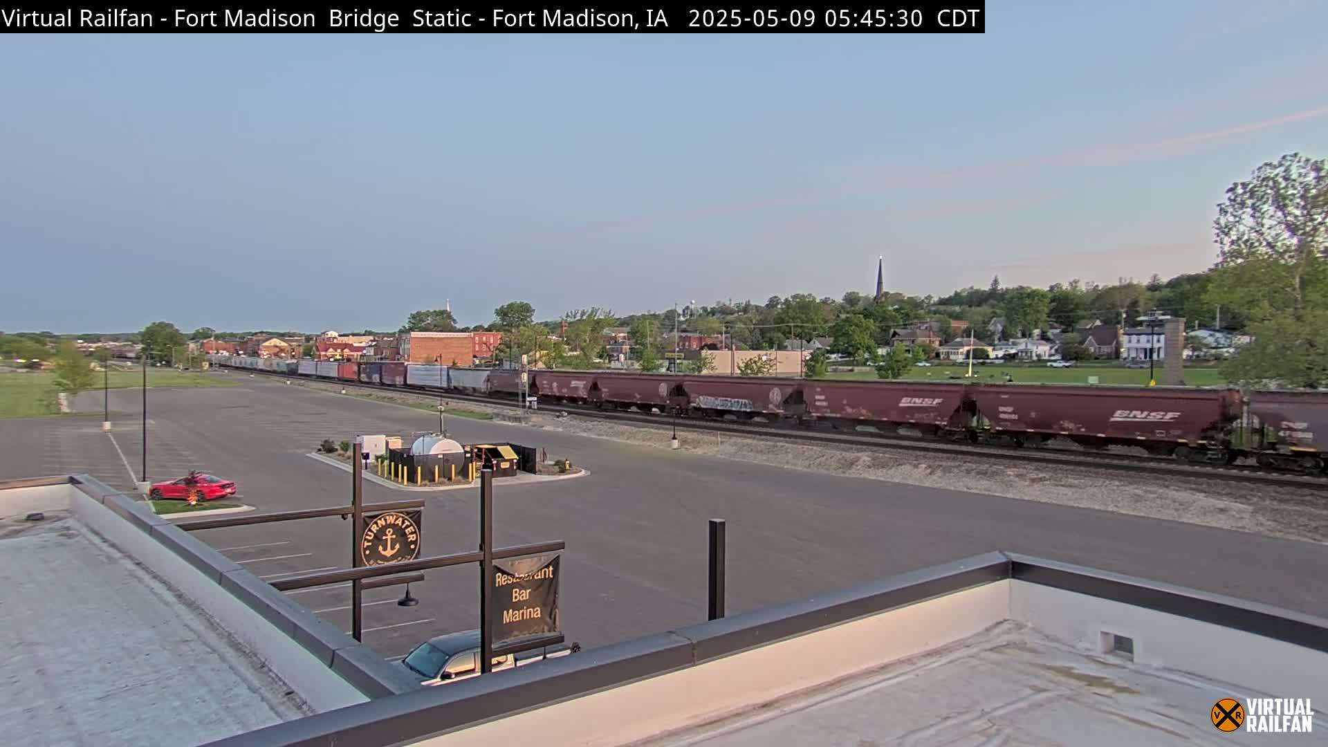 A long freight train travels along railroad tracks in front of a small town under a clear, light-blue sky.