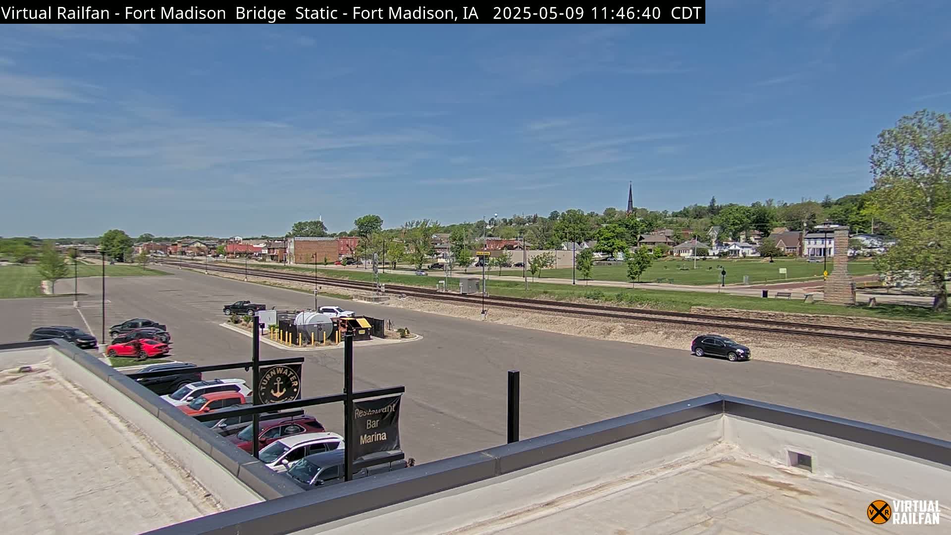 A small town is visible under a mostly clear sky, with a parking lot, railroad tracks, and several cars in view, seen from a rooftop.