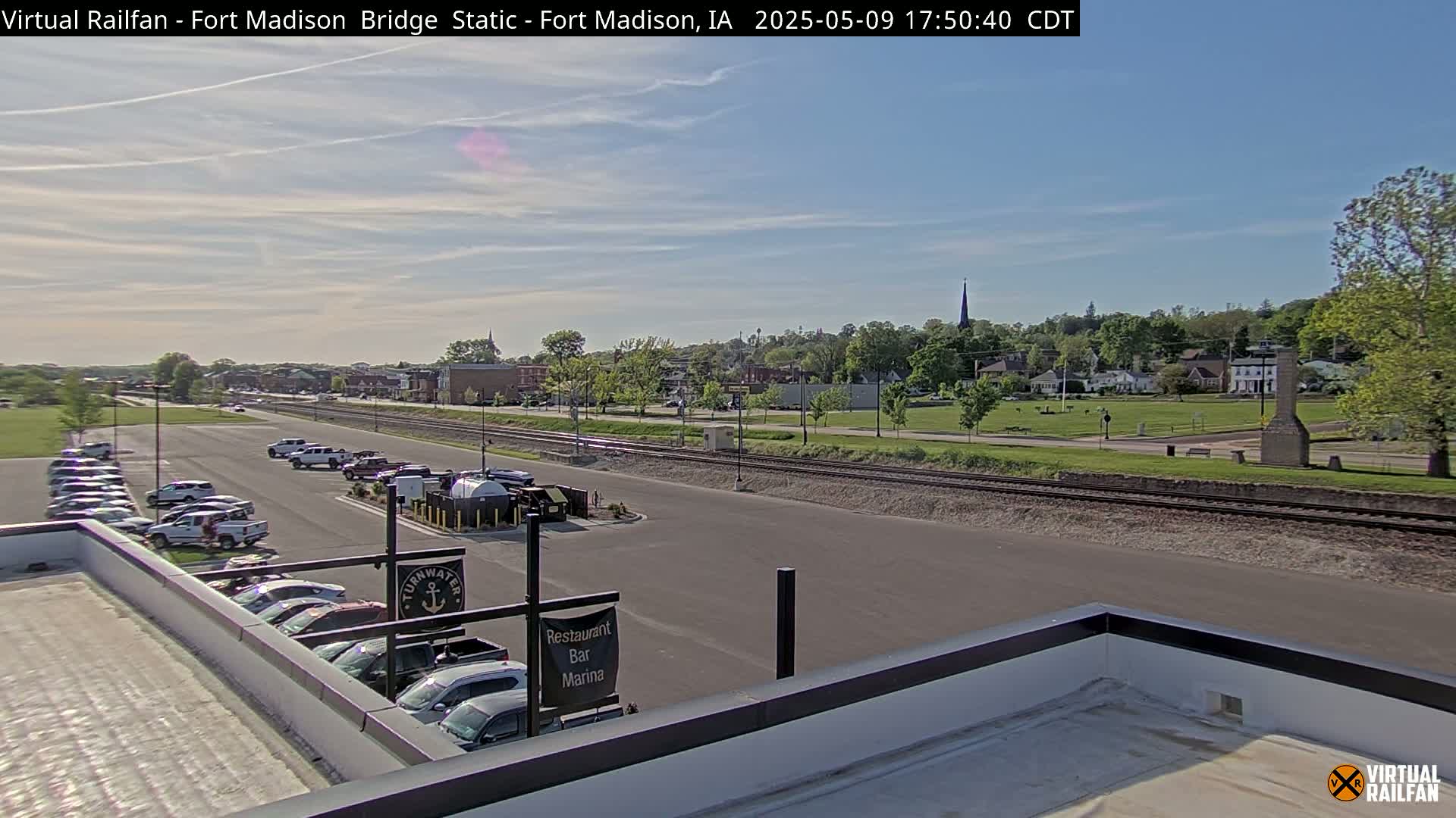 A sunny view of a town with a parking lot and train tracks in the foreground, under a mostly clear sky.