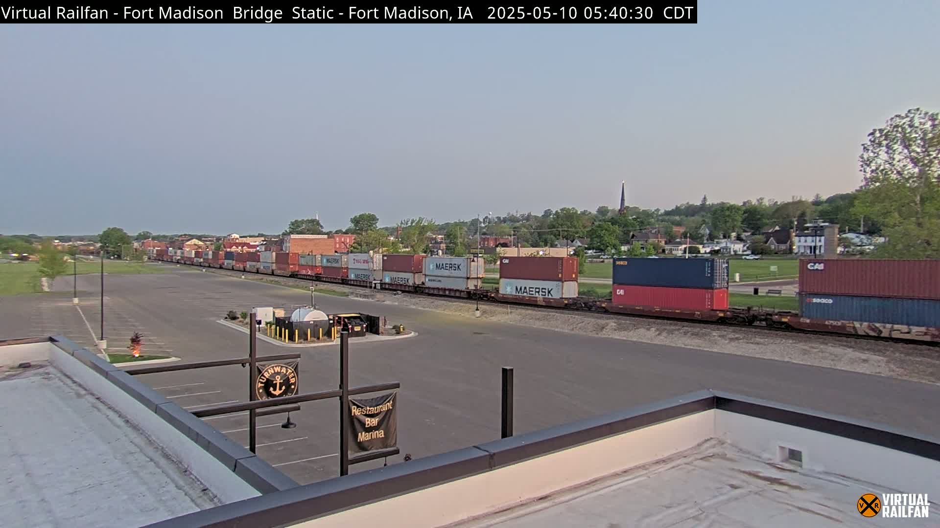 A long freight train carrying multiple shipping containers is parked near a mostly empty parking lot, under a clear, light-grey sky.