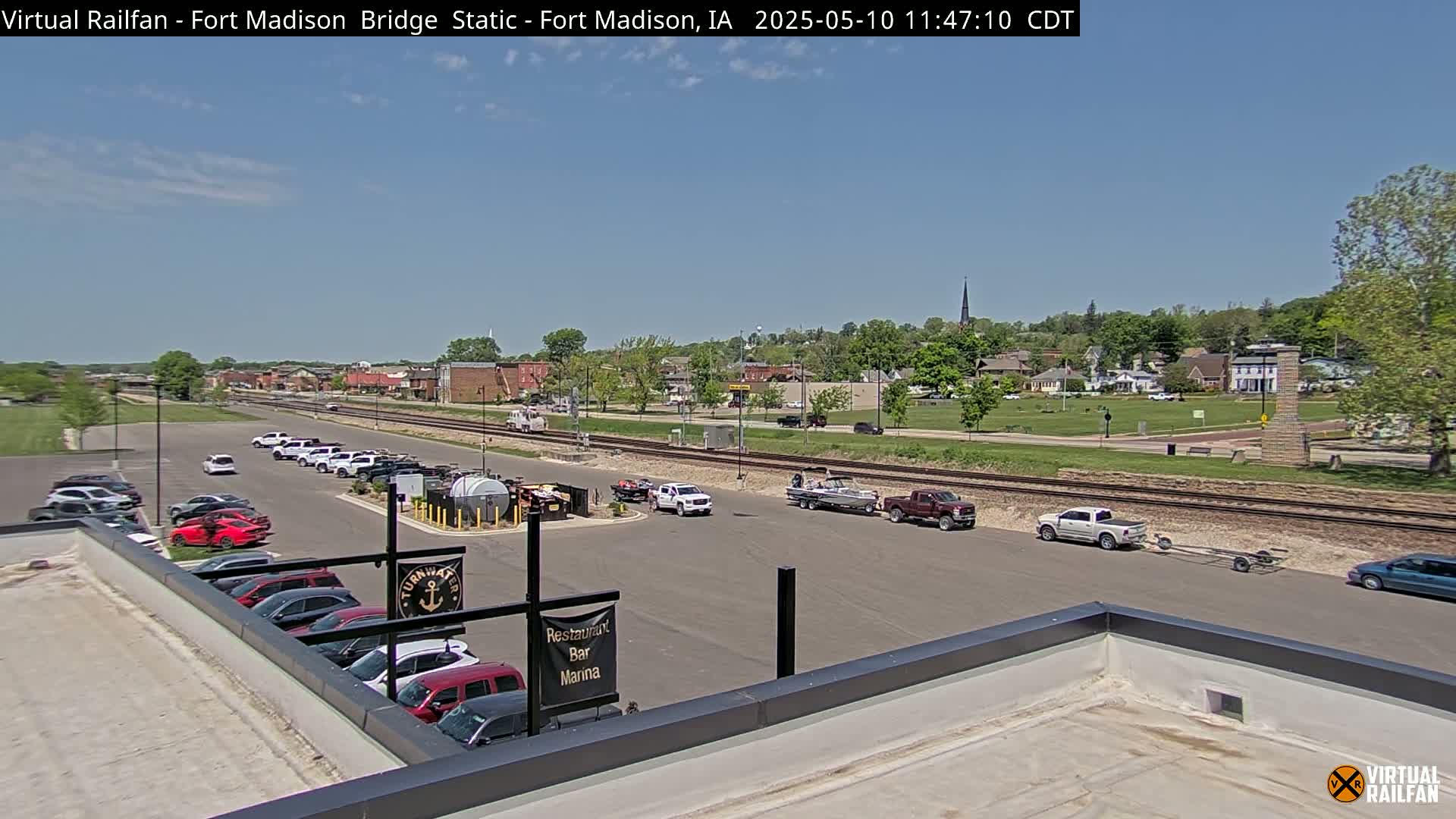 A sunny day shows a small town with a parking lot, cars, trucks, boats, and train tracks, viewed from a rooftop.