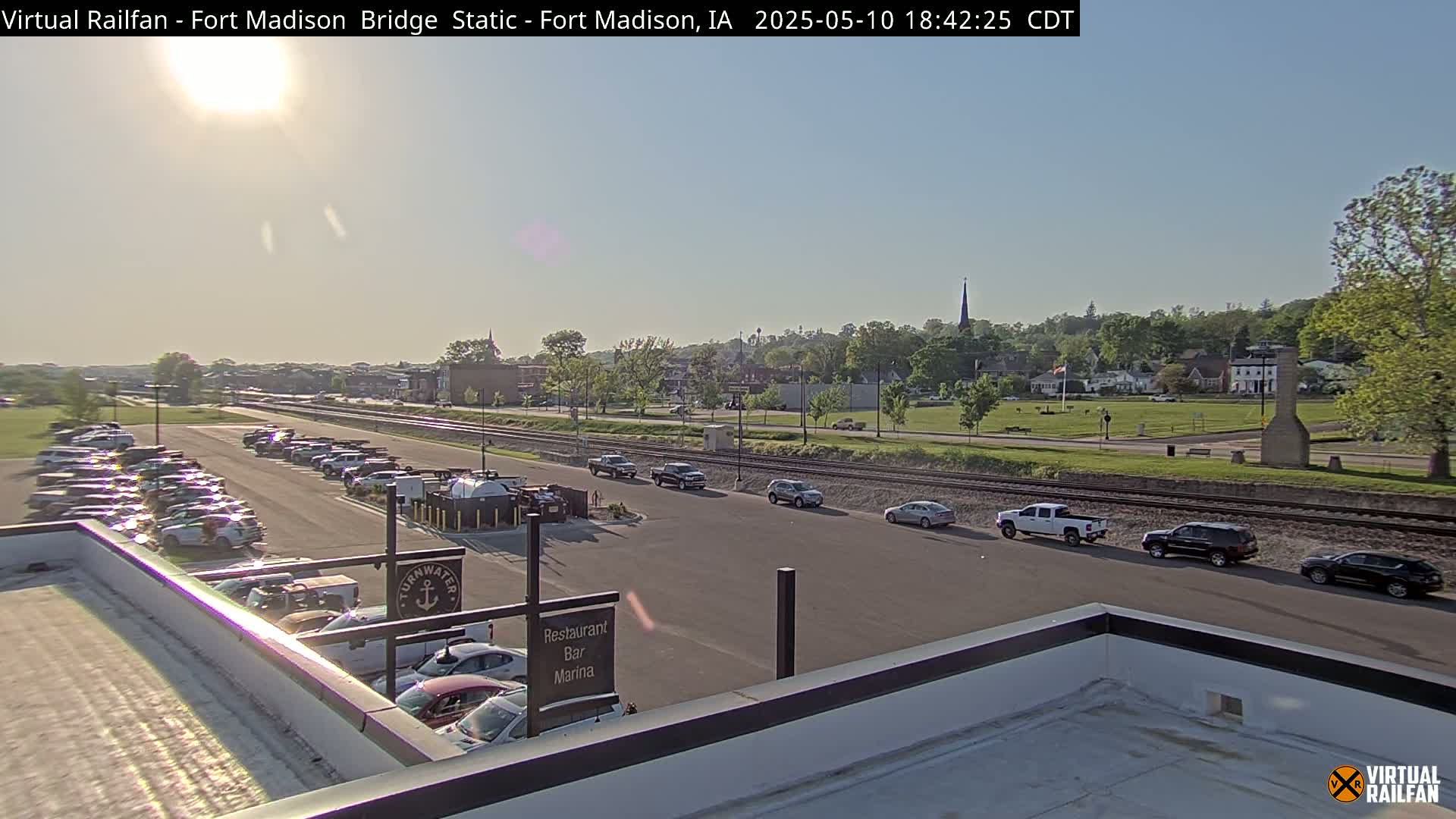 A sunny, daytime view shows a parking lot filled with cars, a railroad track, and a small town in the background.