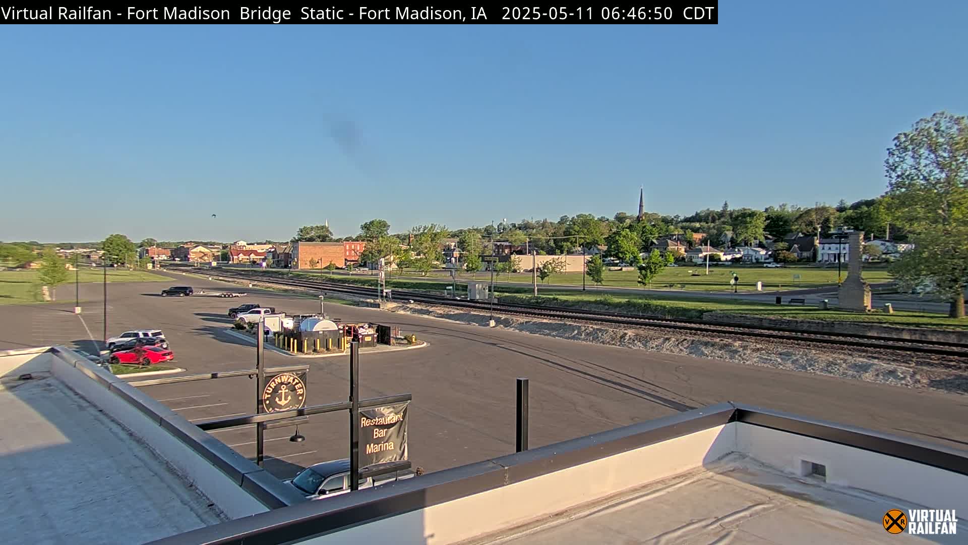 A clear, sunny day reveals a town's cityscape viewed from a rooftop, featuring a parking lot, railroad tracks, and a small body of water in the distance.