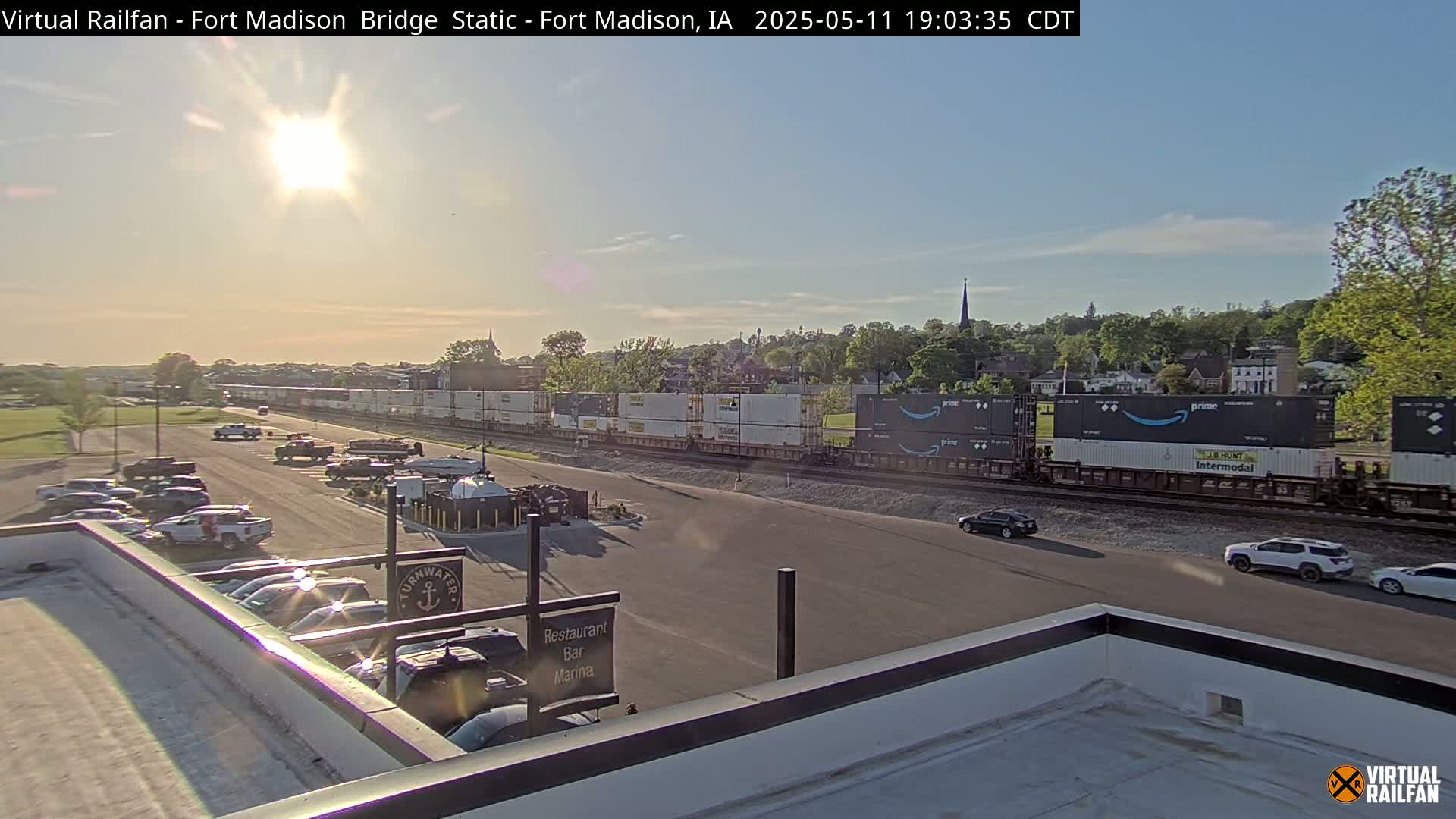 A long freight train passes by a parking lot and restaurant under a bright, sunny sky.