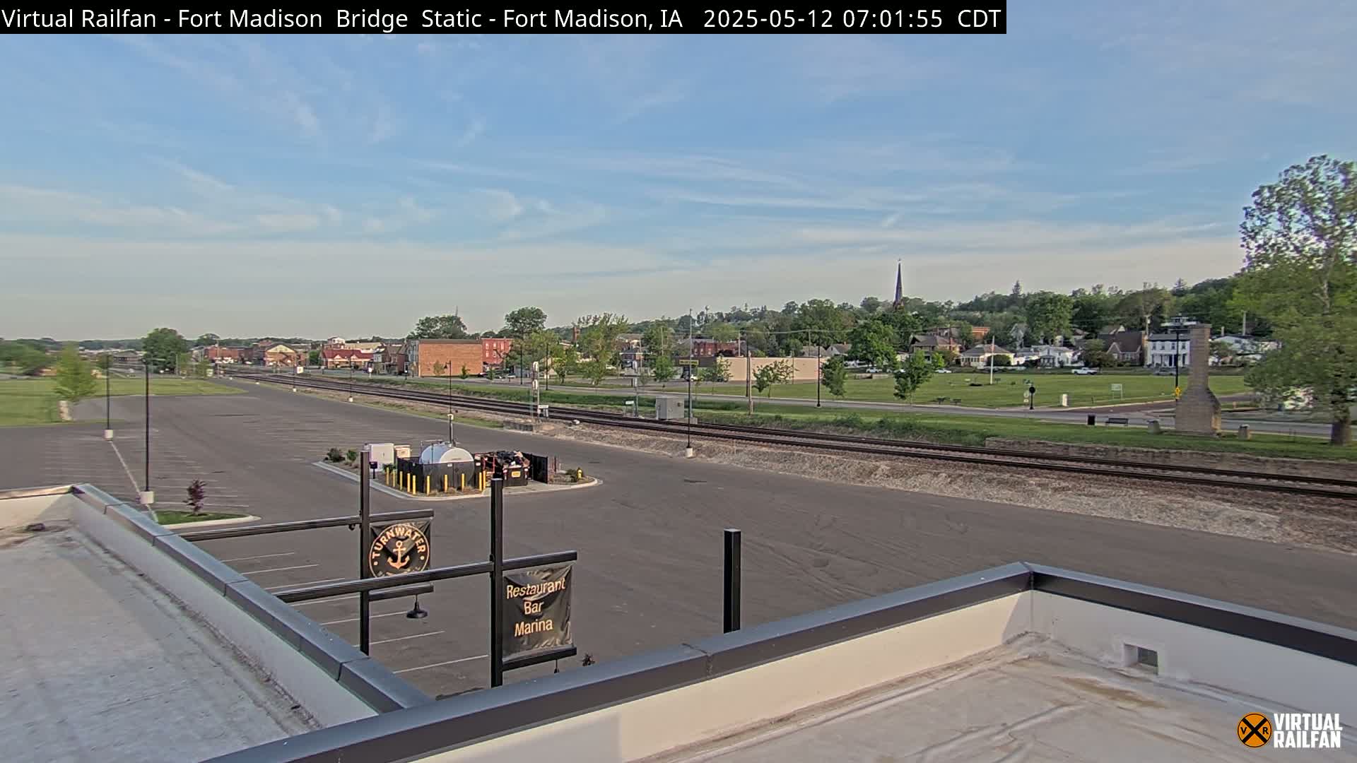 A mostly clear, sunny day reveals a view of a small town across a parking lot and train tracks from a rooftop vantage point.
