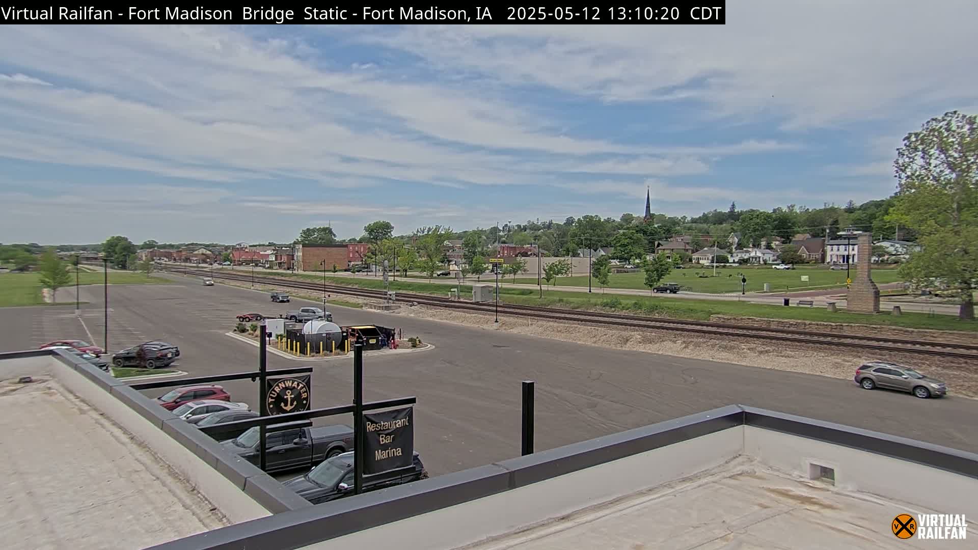 A partly cloudy day reveals a view of a small town, railroad tracks, a parking lot with several cars, and a flat roof with a sign indicating a restaurant, bar, and marina.