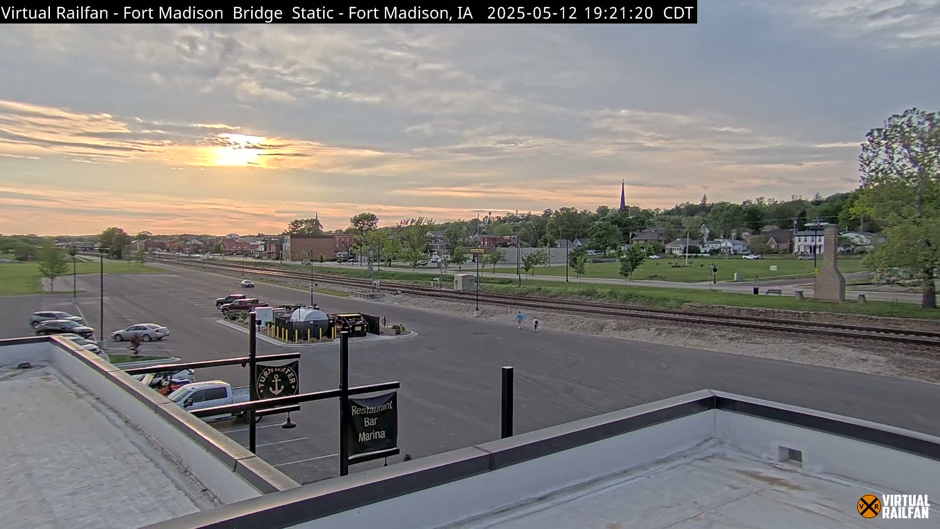 A sunset illuminates a small town with a parking lot, railroad tracks, and a few pedestrians visible in the foreground, viewed from a rooftop.