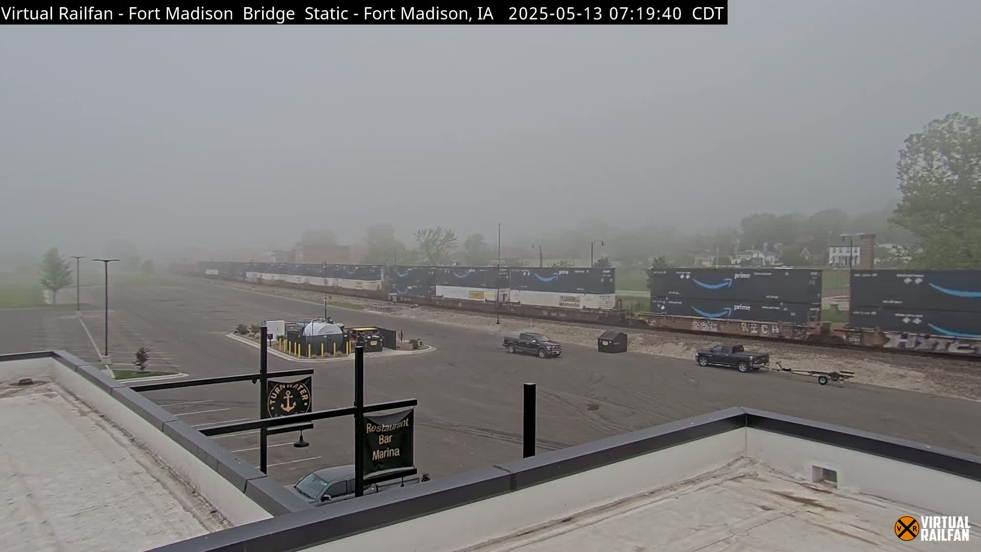 A long freight train carrying Amazon containers sits in a foggy parking lot near a building with a restaurant, bar, and marina.