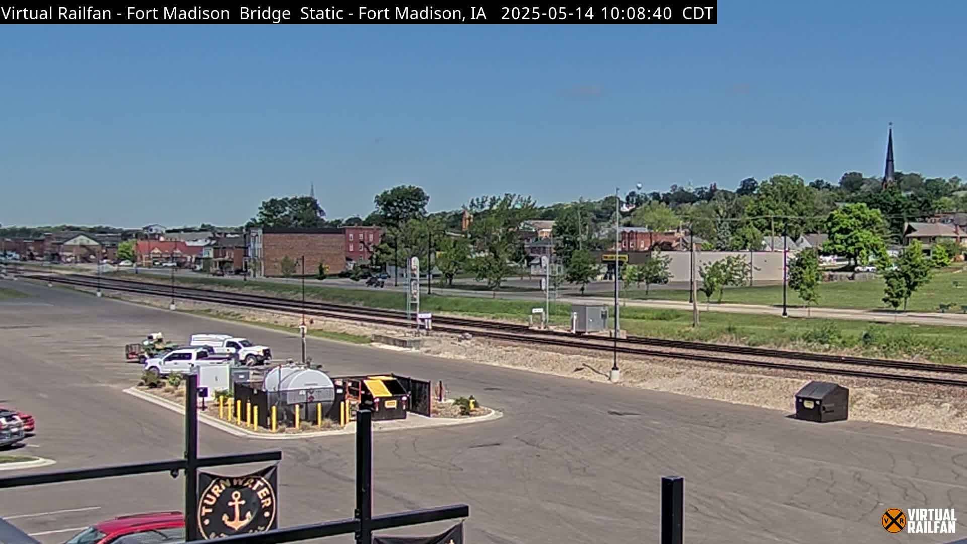 A sunny day shows a parking lot with several vehicles, a railway track, and a small town in the background.