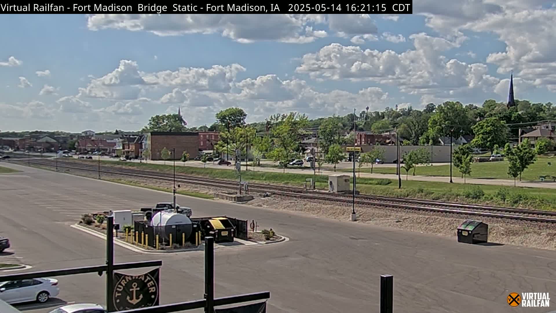 A partly cloudy daytime view shows a small town with railroad tracks in the foreground, a parking lot, and some buildings, including a church steeple.