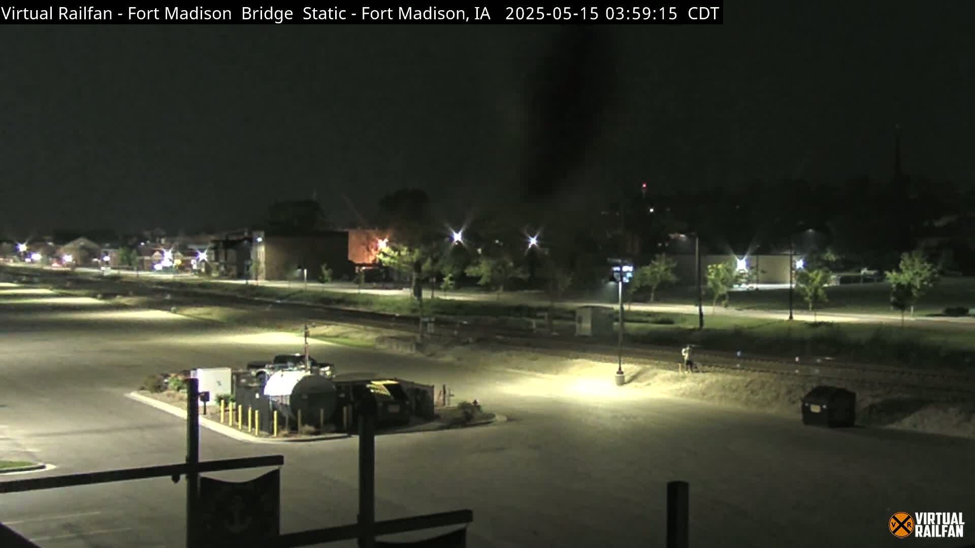A nighttime, well-lit view of an empty parking lot near train tracks and buildings.