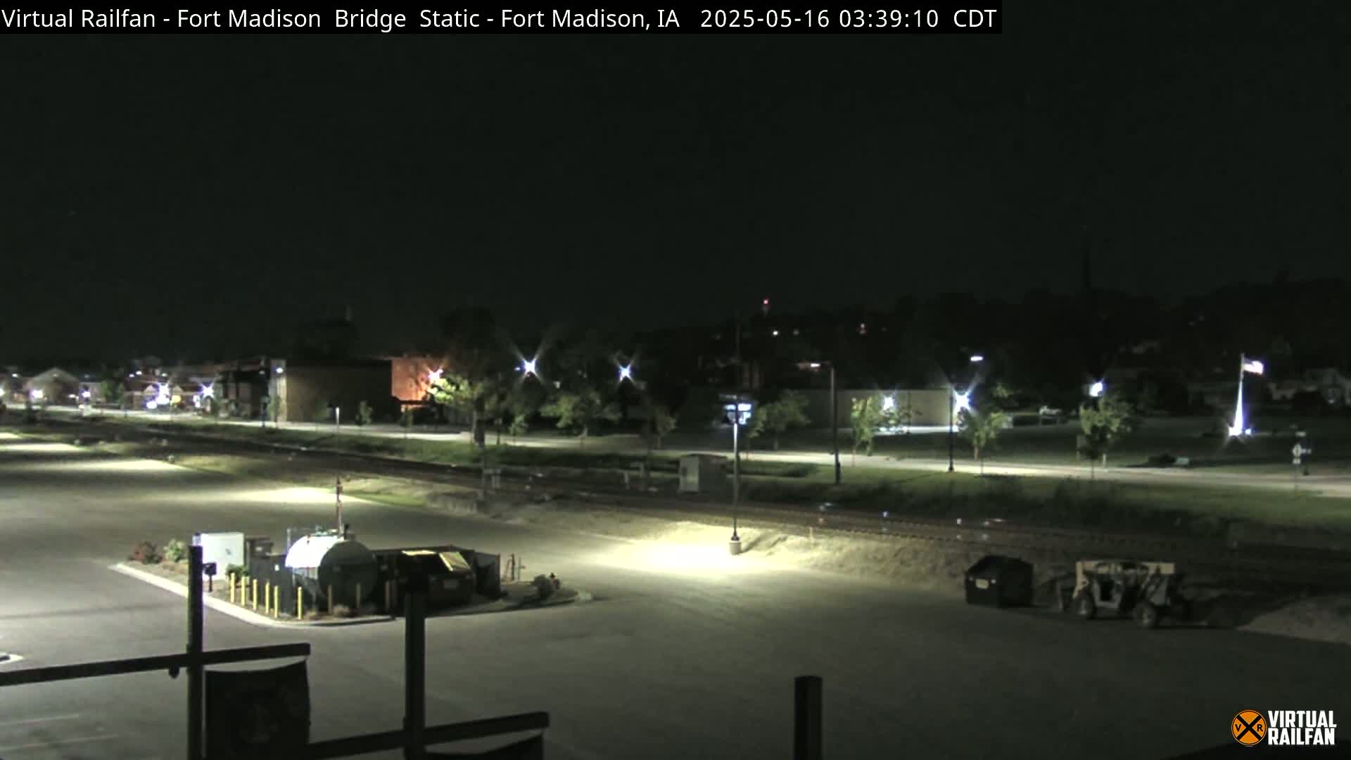 A nighttime view of a mostly empty parking lot next to railroad tracks, with a few vehicles and some buildings visible in the distance under clear skies.