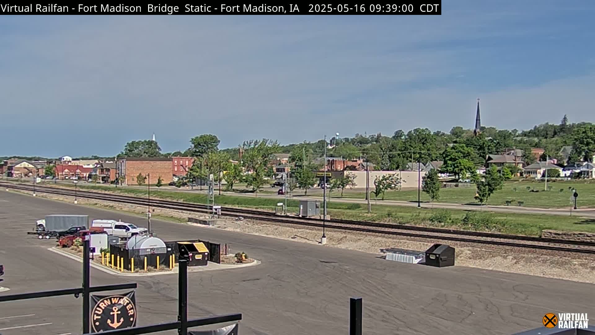 A mostly sunny day shows a small town with train tracks in the foreground and several vehicles parked nearby.