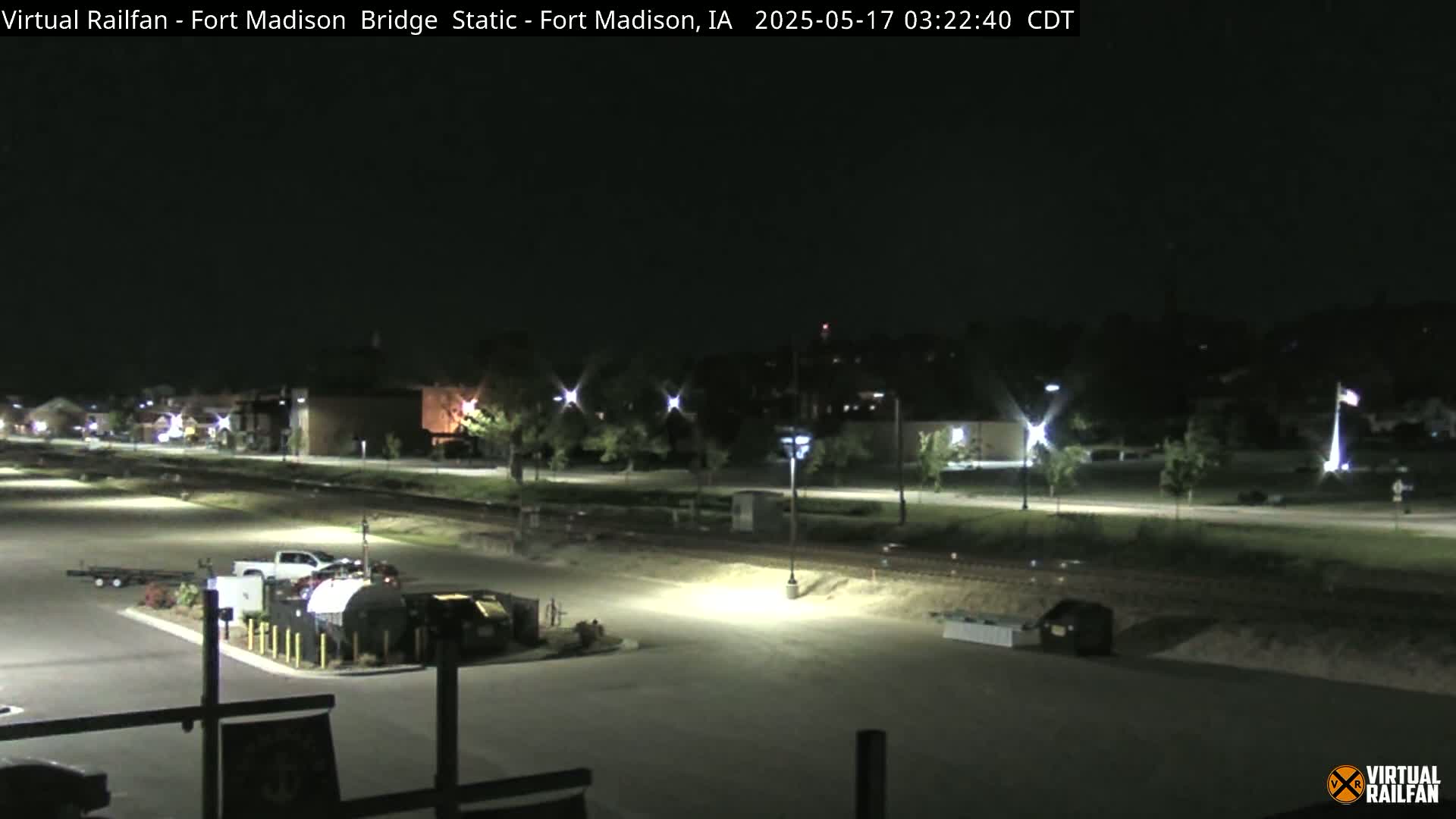 A nighttime, well-lit view of an empty parking lot adjacent to a roadway and railroad tracks, with buildings and trees visible in the distance.