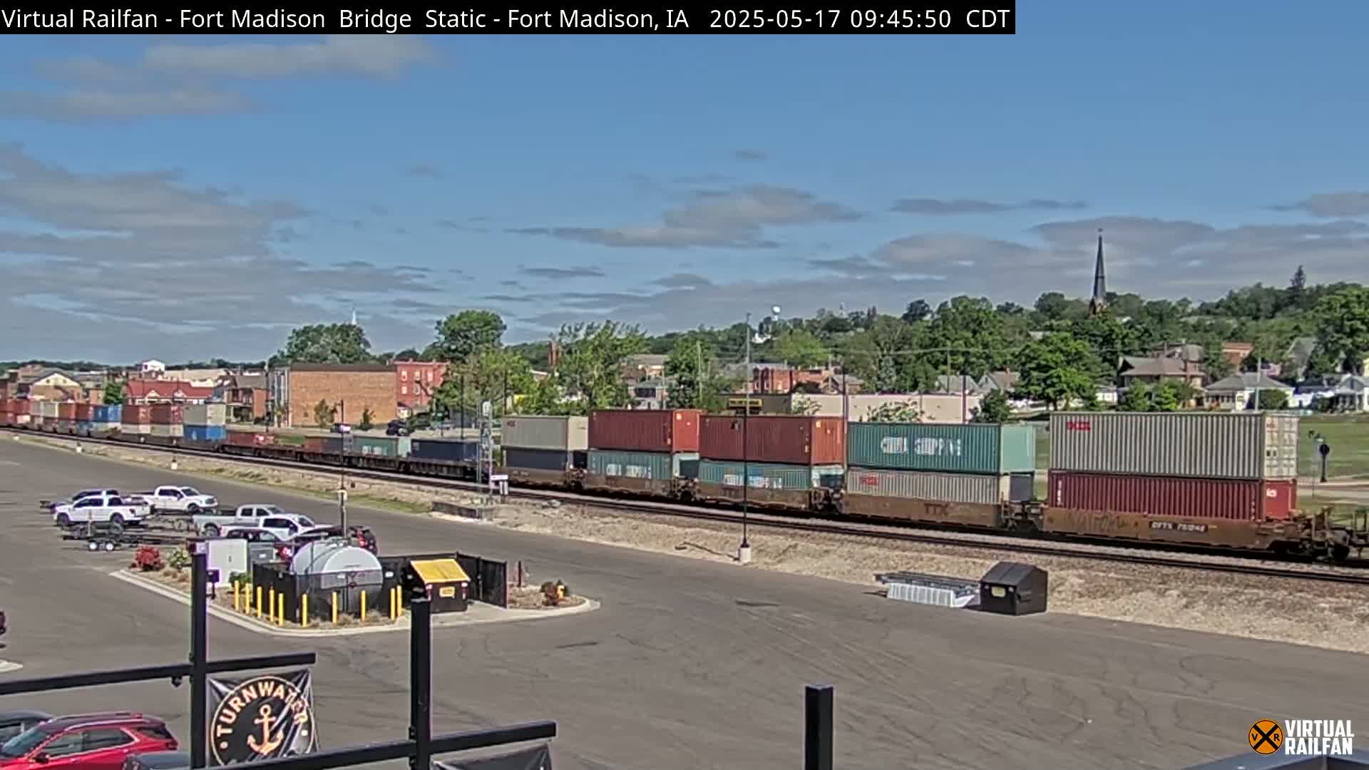 A long freight train carrying stacked shipping containers is moving through a town under a partly cloudy sky.