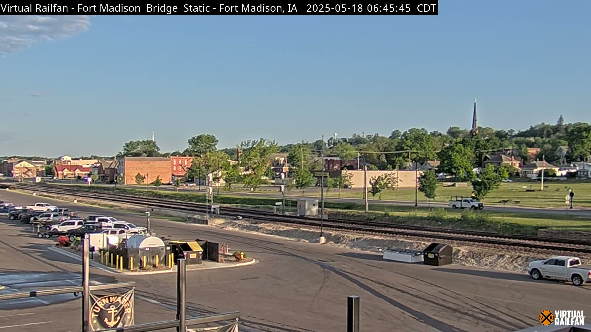 A small town with a parking lot, railroad tracks, and a few vehicles is visible under a clear, sunny sky.
