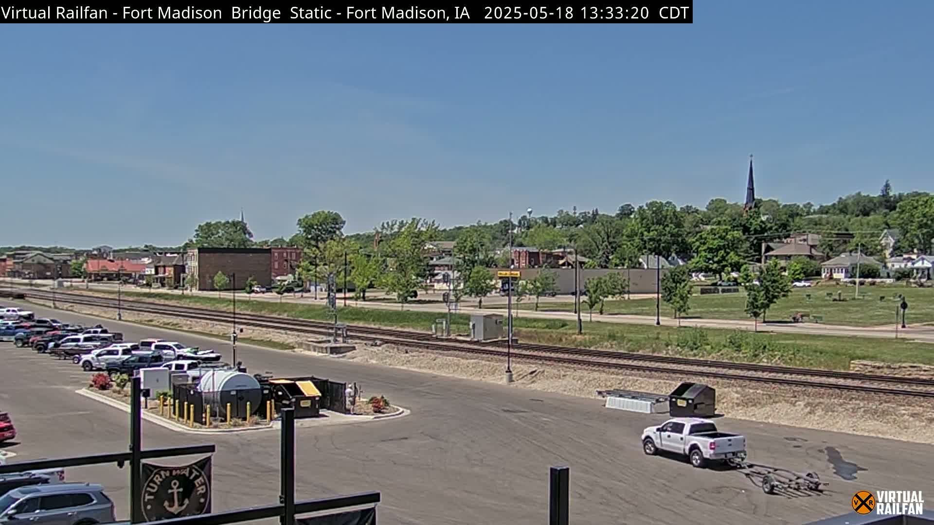 A sunny day reveals a view of a small town across train tracks, with a parking lot, a pickup truck towing a trailer, and some industrial equipment in the foreground.