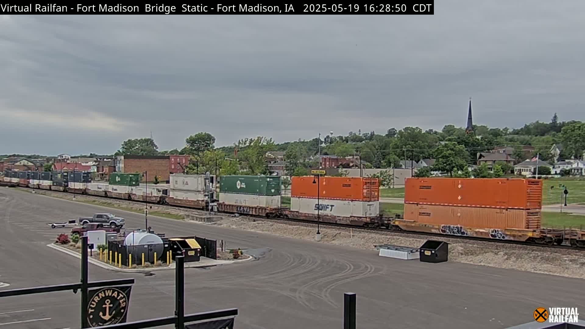 A long freight train with various colored shipping containers passes under an overcast sky next to a parking lot.