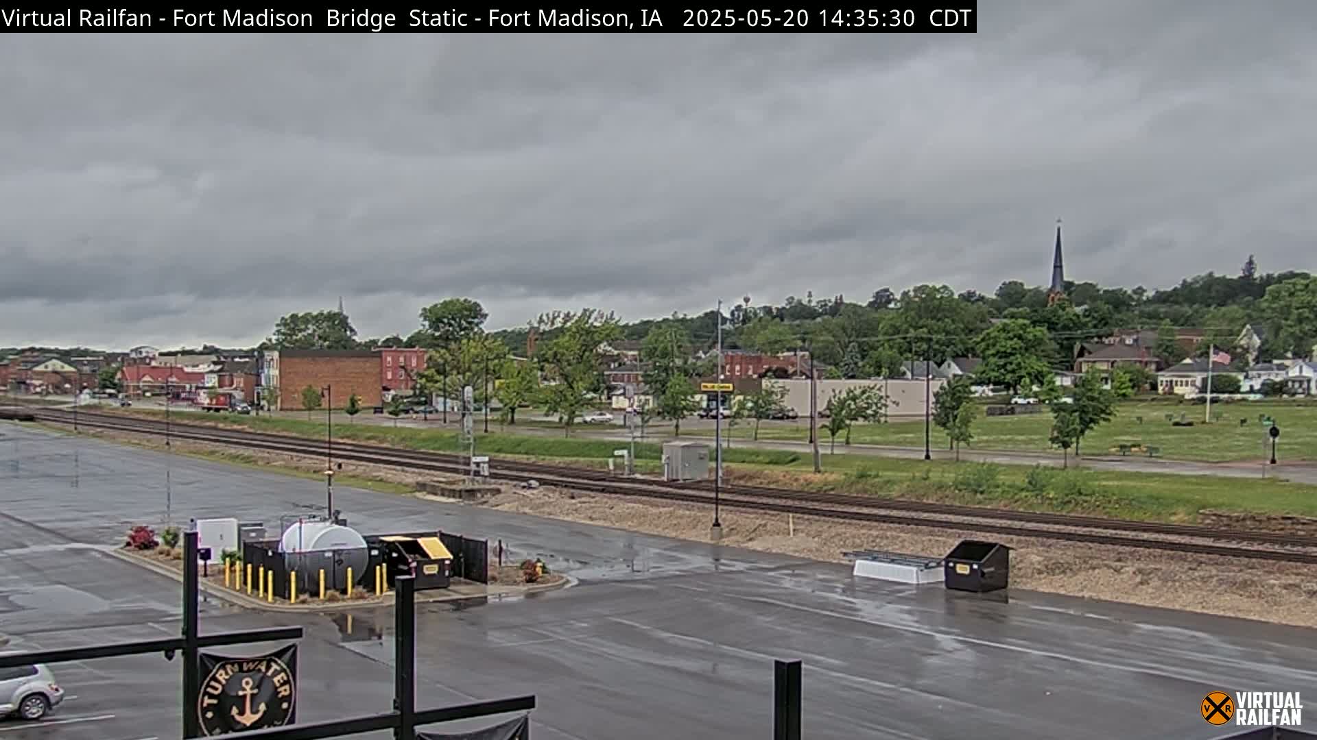 A wet, overcast parking lot overlooks railroad tracks and a small town under a cloudy sky.