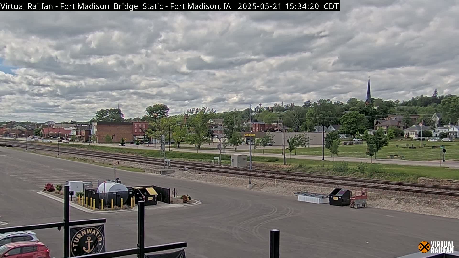 A view of a town under a partly cloudy sky, showing a parking lot, railroad tracks, and a small collection of buildings beyond.