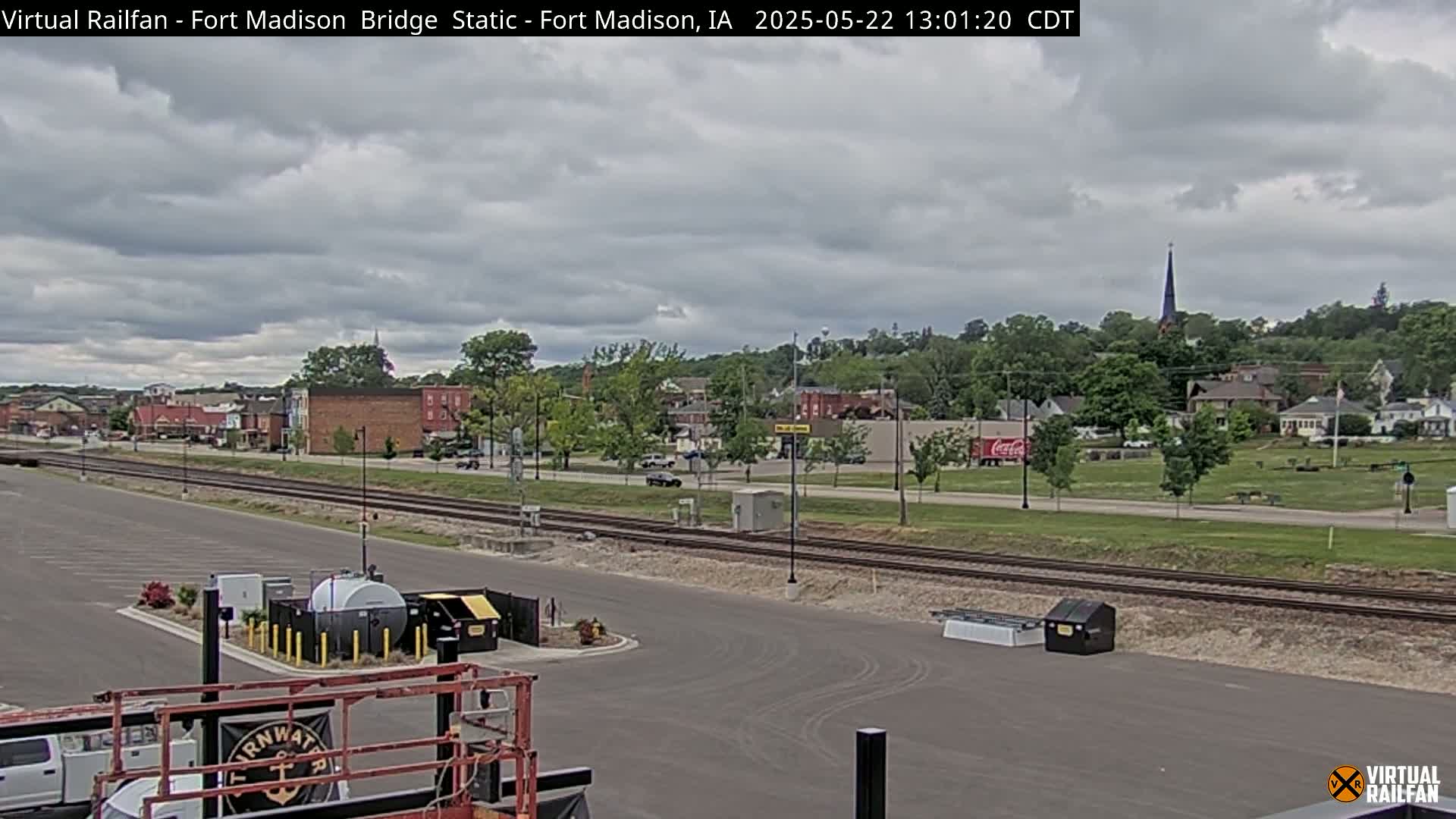 A mostly cloudy day reveals a small town with railroad tracks in the foreground and a church steeple visible in the distance.