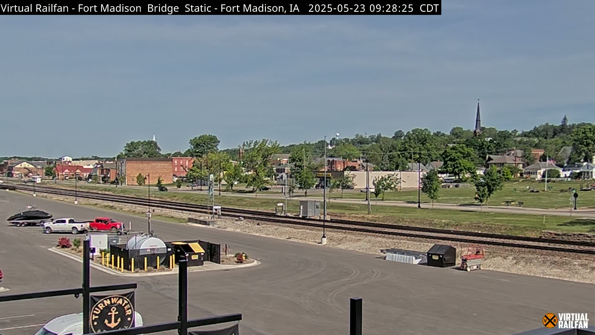 A mostly sunny view of a town with railroad tracks in the foreground and a church steeple visible in the distance.