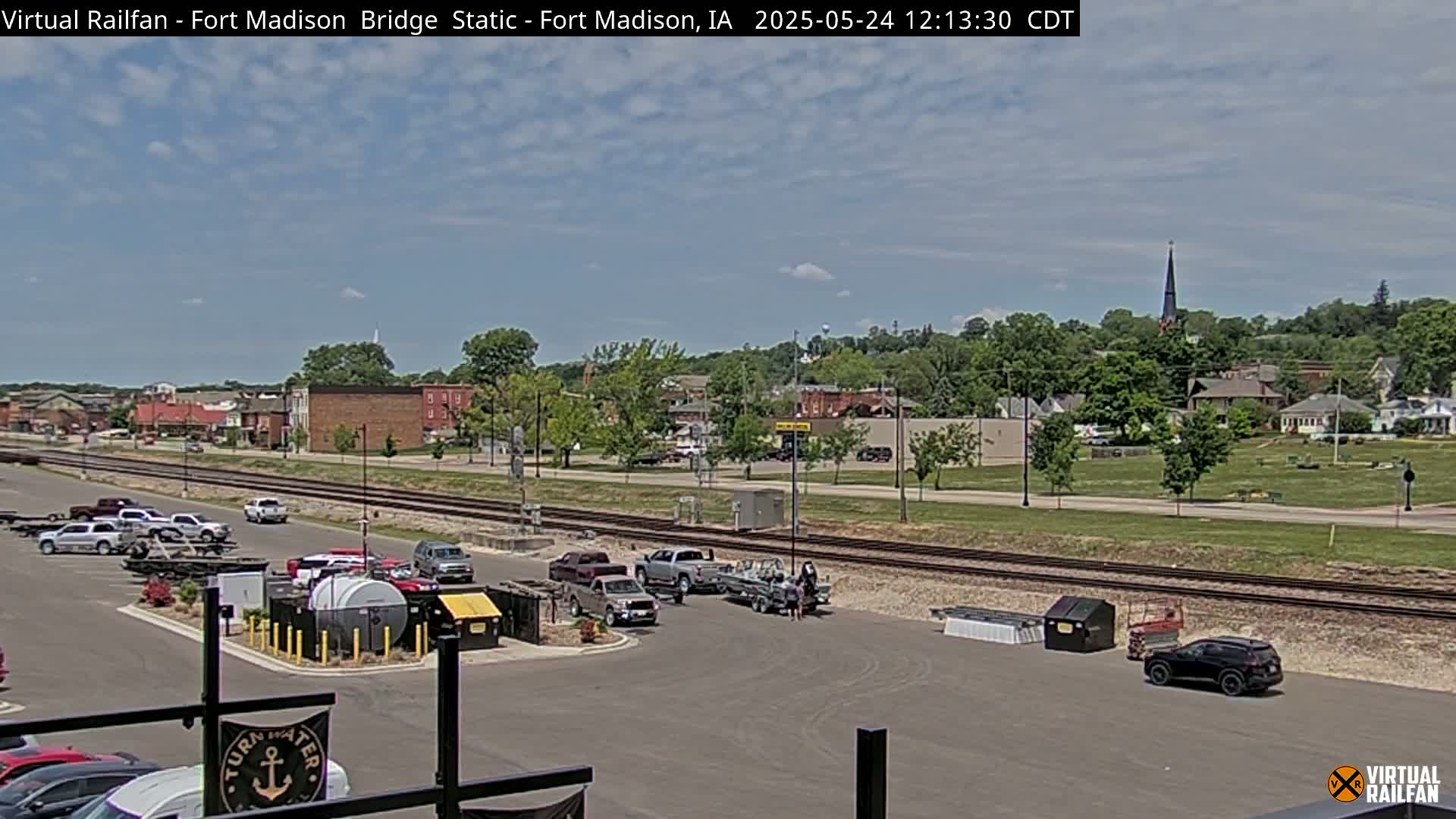 A partly sunny day shows a parking lot with several vehicles, boats, and trailers near train tracks, overlooking a small town with a church steeple visible in the distance.