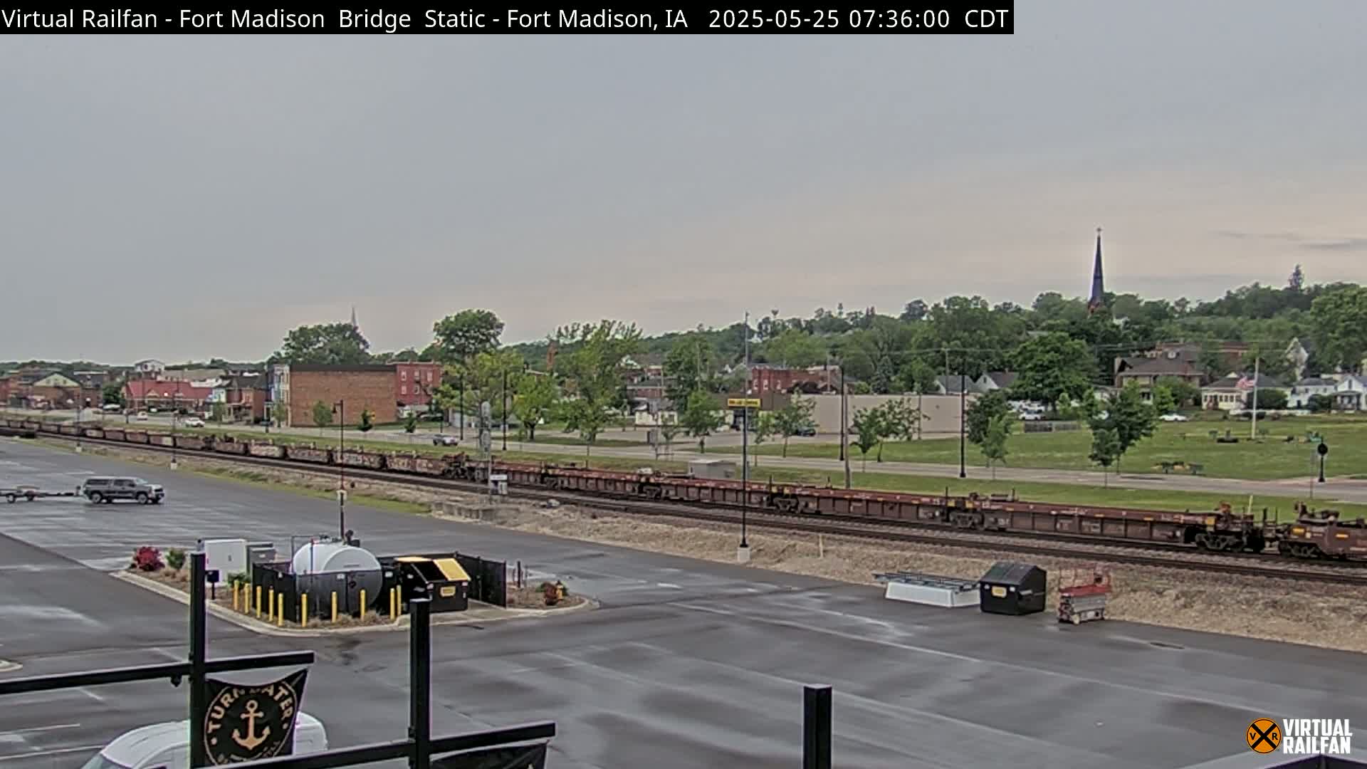 A long freight train sits on railroad tracks in a mostly empty parking lot, with a small town visible in the background under an overcast sky.
