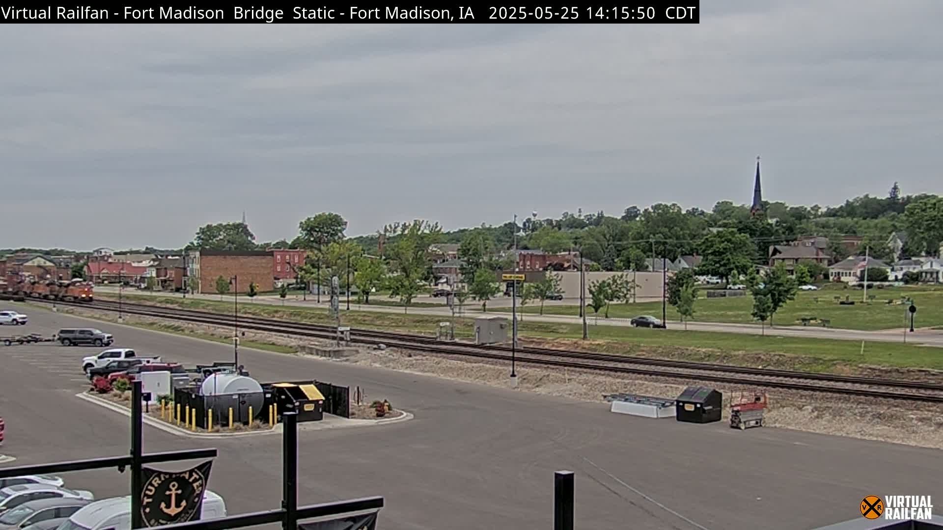 A long freight train passes by a small town under an overcast sky, with a parking lot and railroad tracks visible in the foreground.