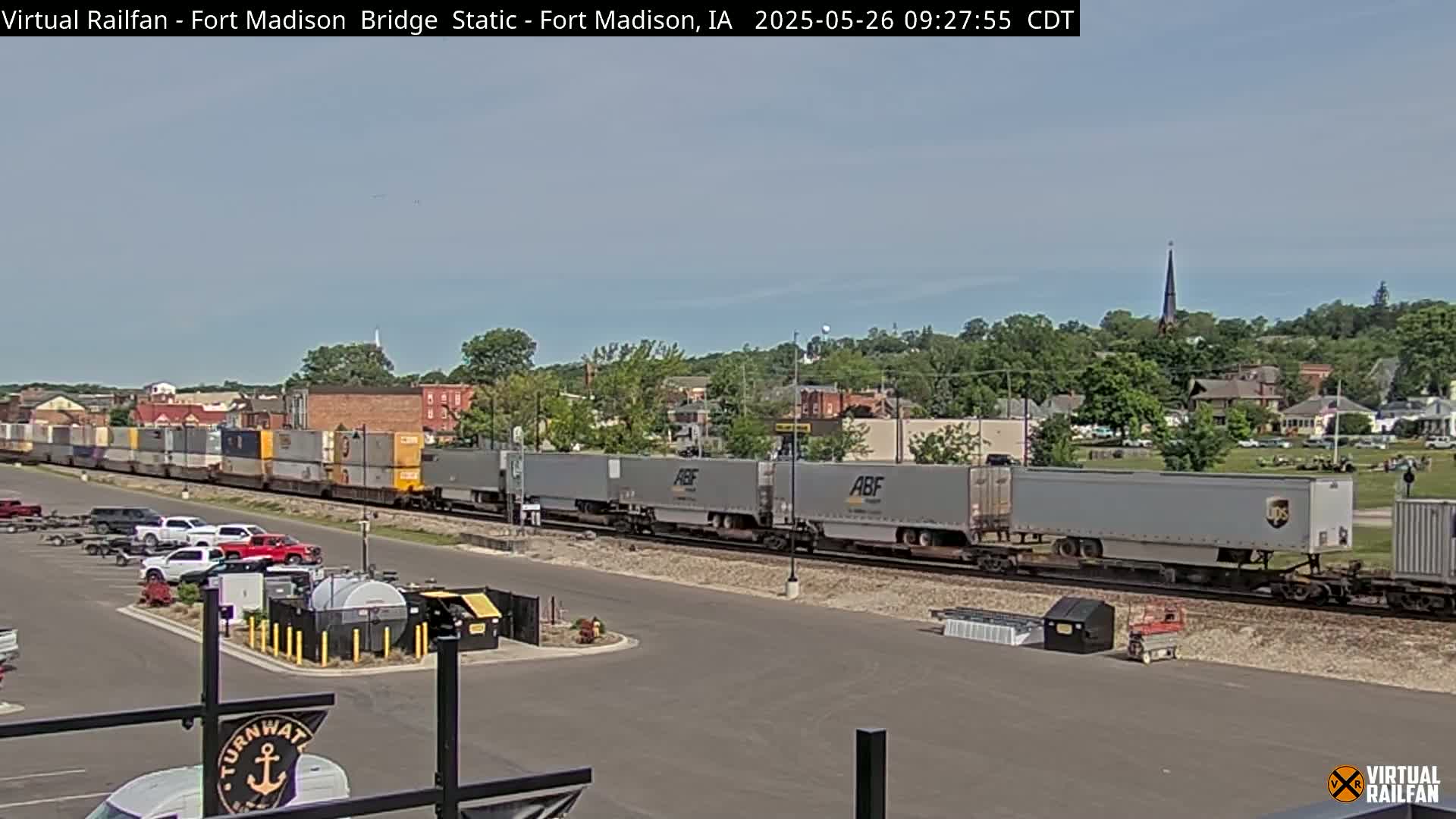 A long freight train passes through a town on a sunny day, with several cars parked nearby.