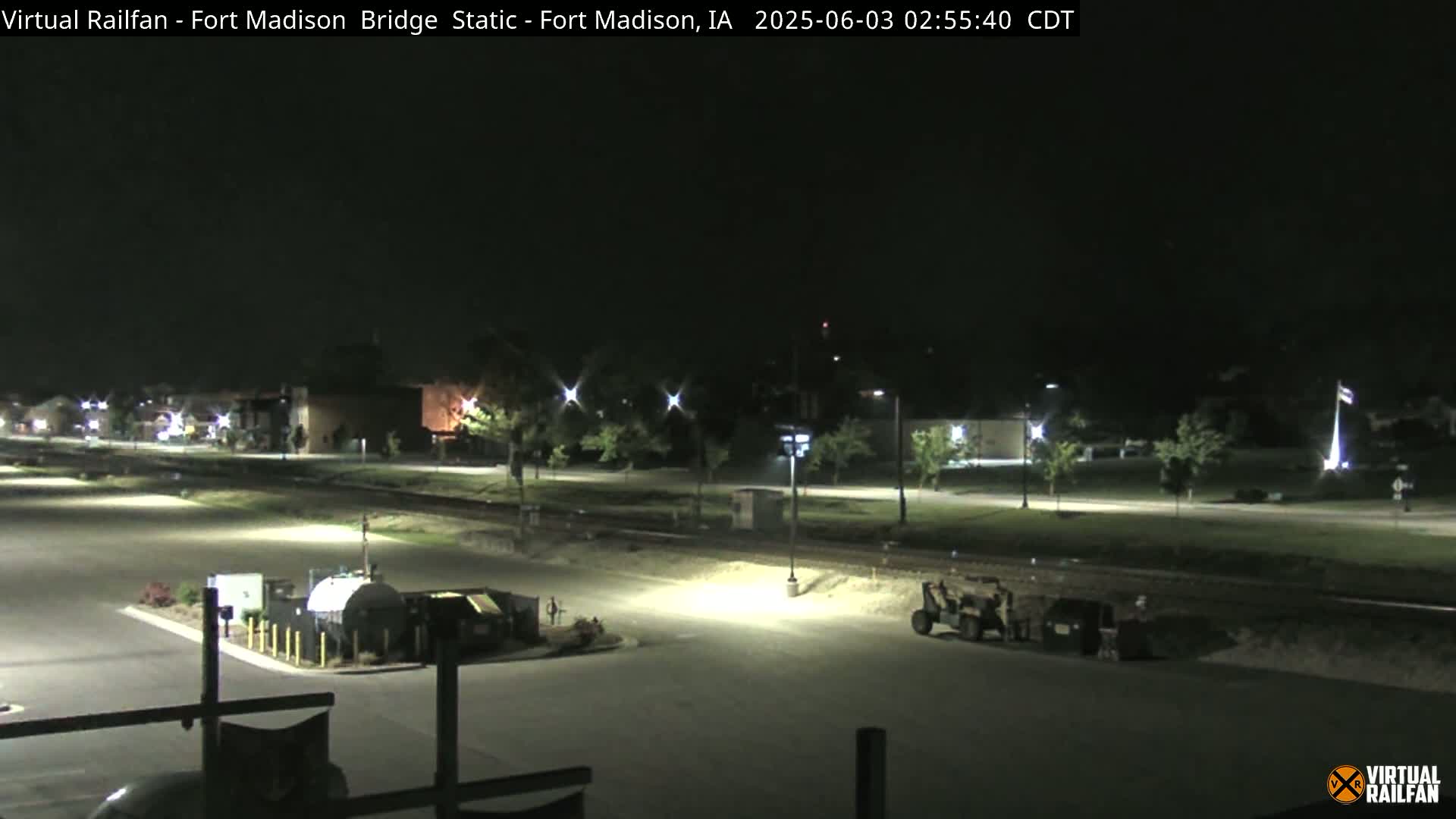 A nighttime view of an empty parking lot and roadway, illuminated by artificial lights, with some trees and buildings visible in the background.