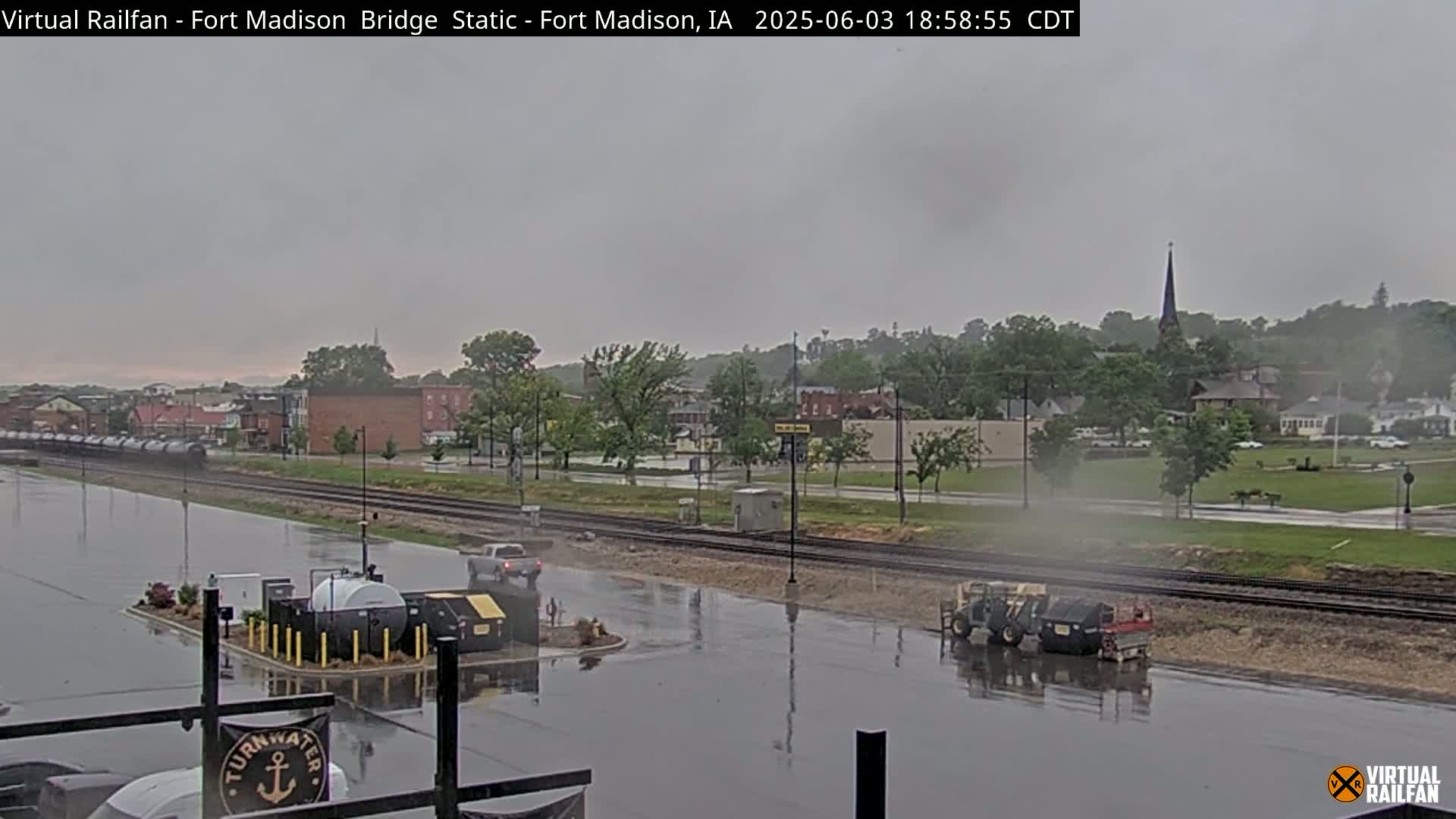 A rain-soaked parking lot and railroad tracks are seen in front of a town under a gray sky, with a few vehicles present.