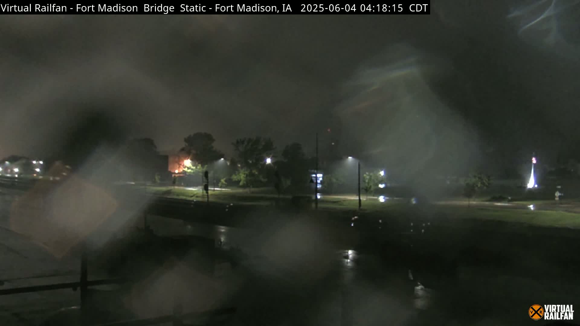 A nighttime view of a rain-soaked street scene, showing a bridge and trees under a dark, stormy sky.