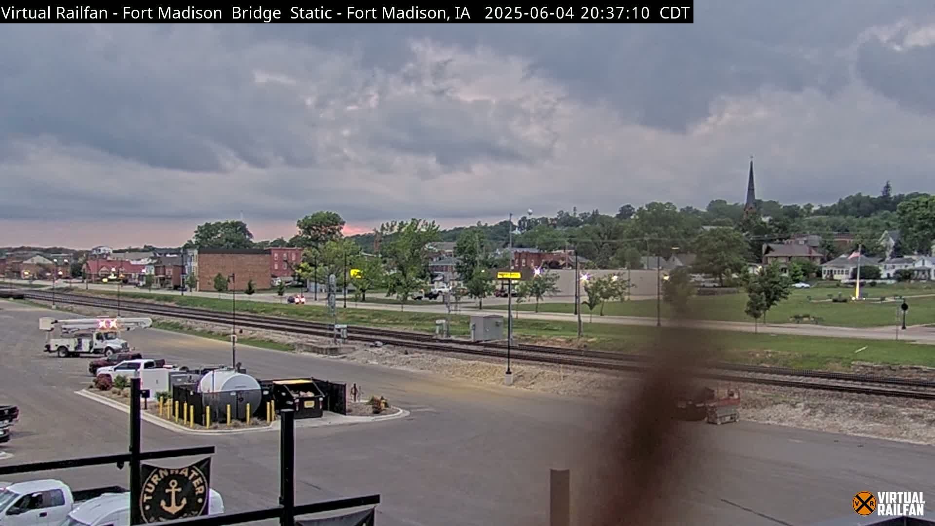 A mostly cloudy evening scene shows a small town with railroad tracks, vehicles in a parking lot, and a church steeple visible in the distance.
