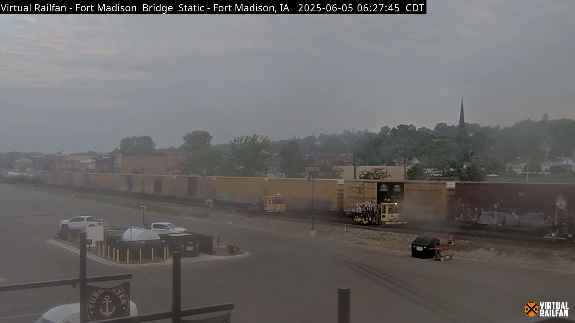 A long freight train is visible in the foreground, with several vehicles parked in a lot, all under a hazy, overcast sky.