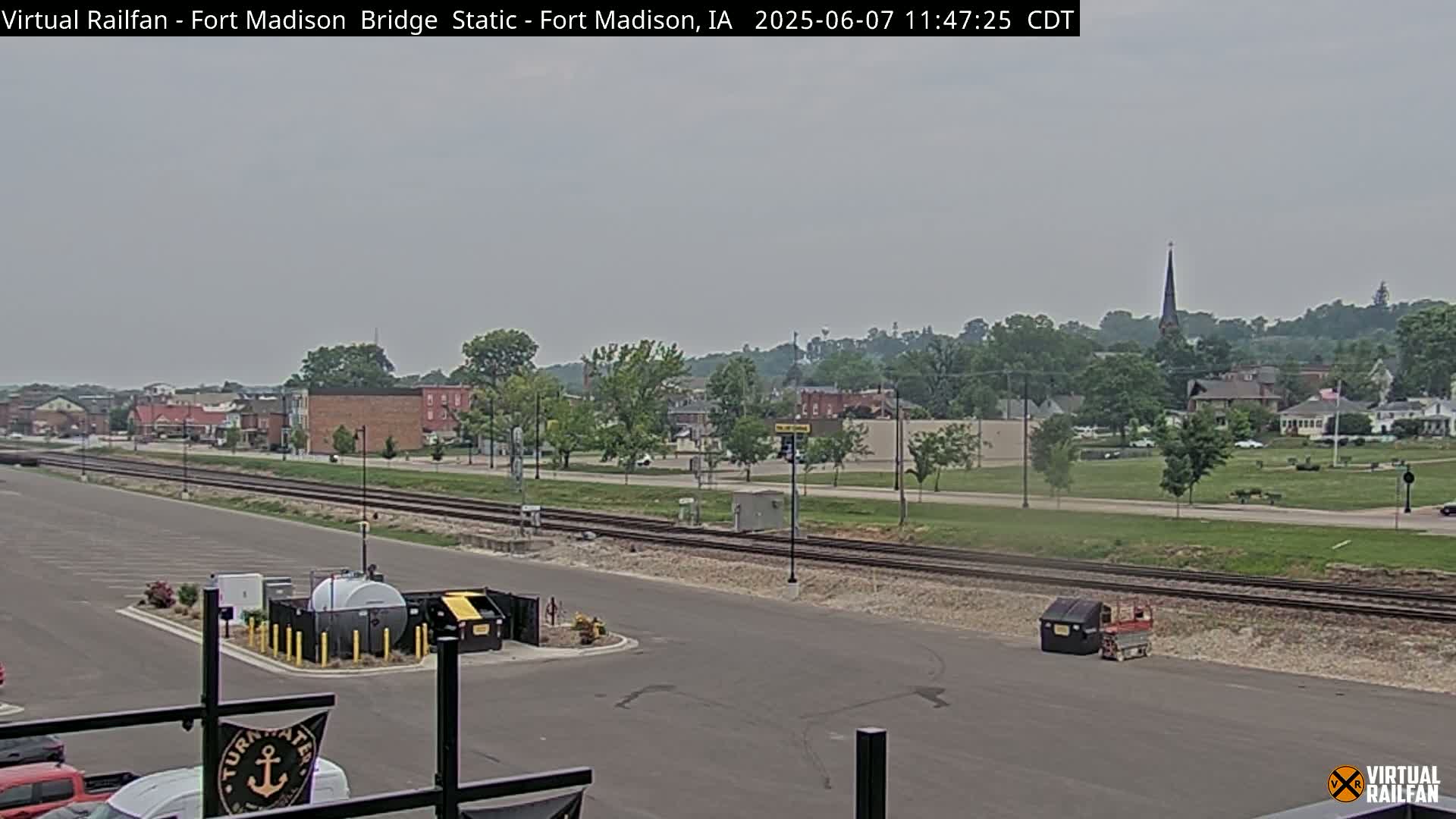 A mostly cloudy day reveals a small town with railroad tracks in the foreground, and a church steeple visible in the distance.
