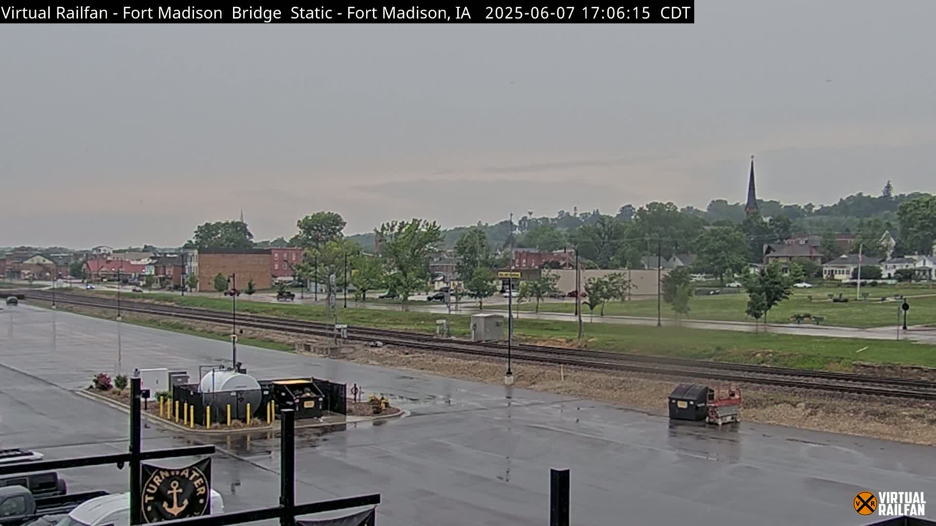 A wet, overcast view of a town with railroad tracks and buildings in the distance, seen from a parking lot.