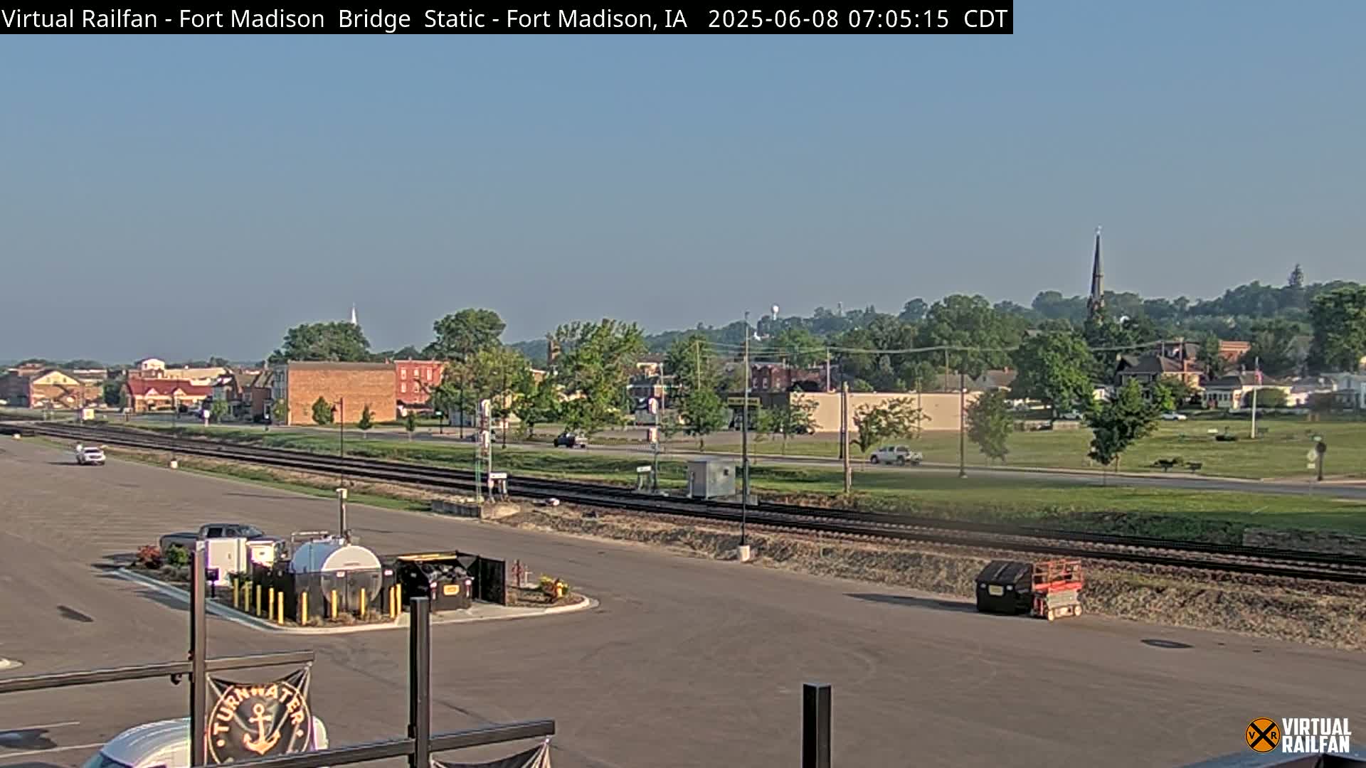 A mostly sunny view of a town, featuring railroad tracks, a parking lot with some vehicles, and a church steeple visible in the distance.