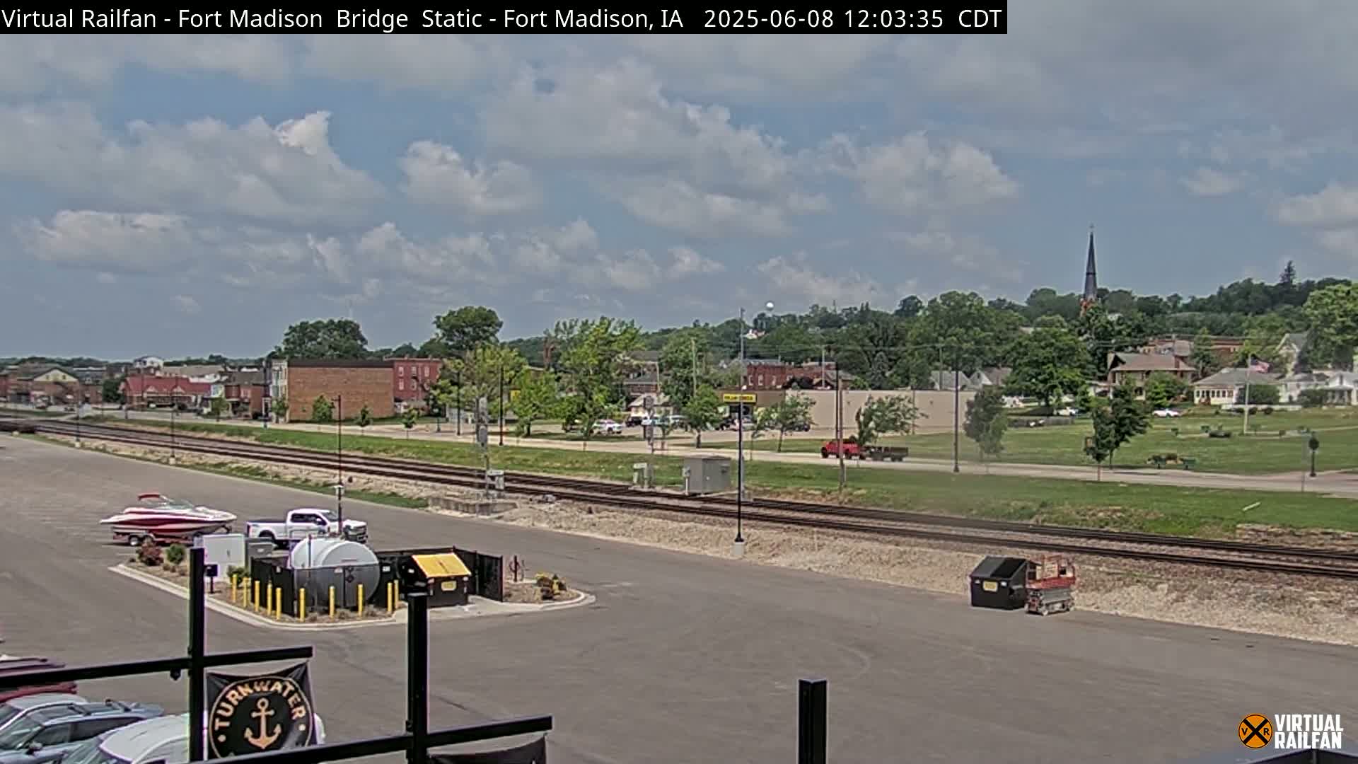 A partly cloudy day reveals a small town with railroad tracks and a boat parked near a parking lot, and some other vehicles nearby.